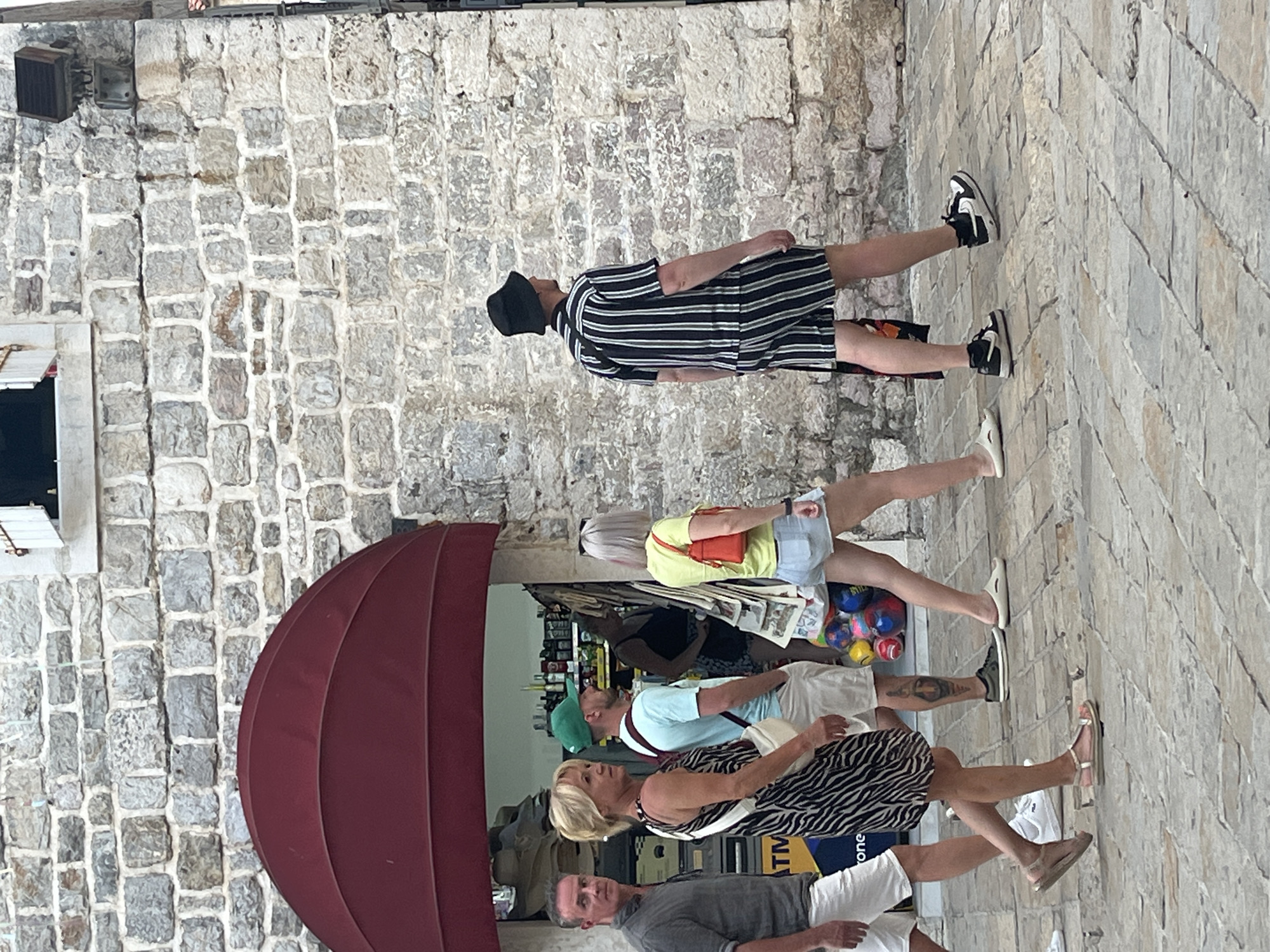 A group of five individuals is walking on a paved street in Budva, Montenegro, next to a stone building with a red awning. On the left, a man in a grey short-sleeved shirt and white shorts walks facing forward. Next to him, a woman in a black and white striped dress walks ahead, with a white strap across her body. Behind her, a man wearing a light blue t-shirt, white shorts, and a green baseball cap walks. To his right, a woman in a yellow short-sleeved shirt and light blue denim shorts carries an orange purse. On the far right, a man in a black bucket hat, a black and white striped short-sleeved shirt, and matching shorts walks facing away from the camera. The background includes a store entrance with various items displayed.