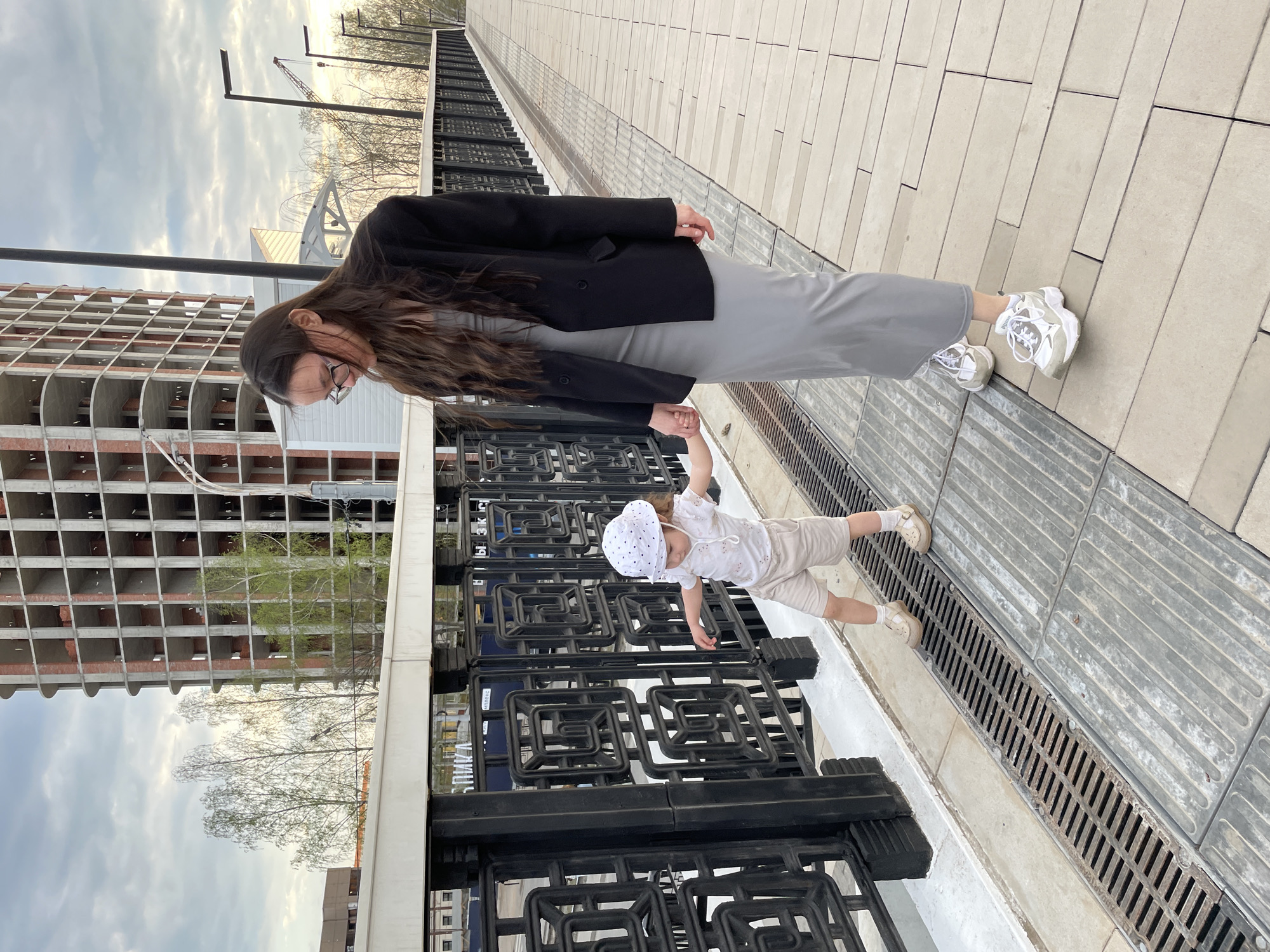 A mother and child stand on a bridge in Izhevsk, Russia. The mother, with long brown hair and wearing glasses, is clad in a grey dress and a black blazer, paired with white sneakers. She looks down at the child, a toddler wearing a patterned white hat, shorts, and light-colored shoes, who is grasping the bridge's black metal railing. In the background is an unfinished, multi-story building, revealing its brick structure. The sky is overcast, and the bridge's surface is paved with light-colored tiles. A drainage grate runs along the edge of the bridge.