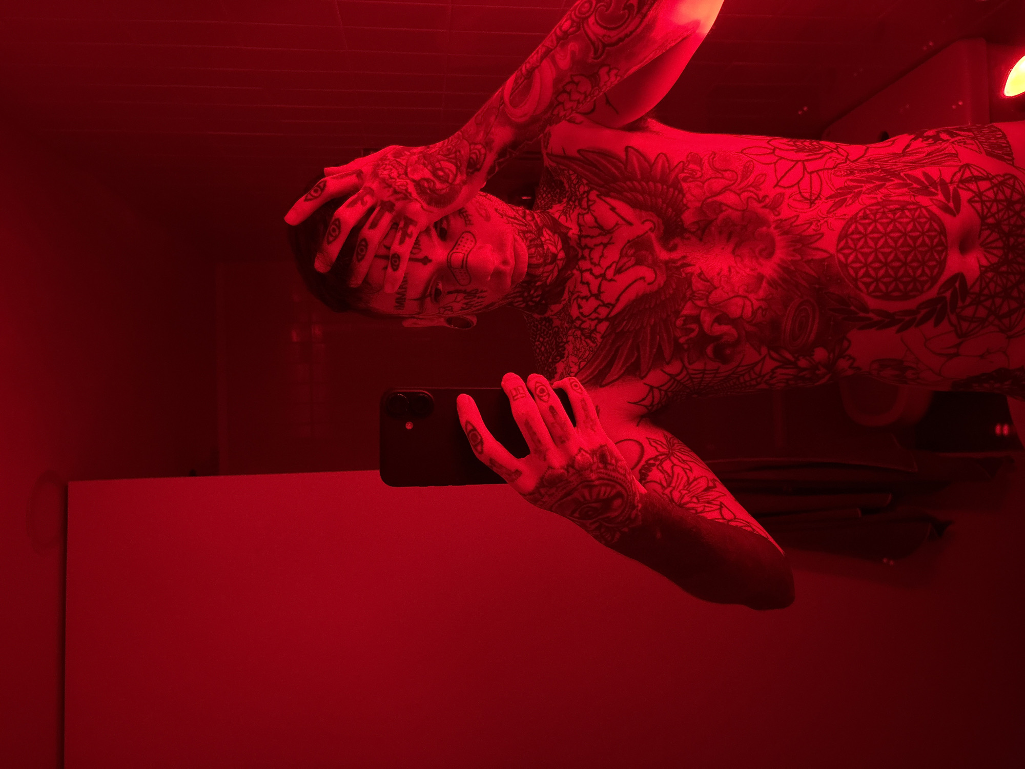 A partially nude, heavily tattooed person is reflected in a mirror in a red-lit room. The person is holding a black smartphone and appears to be taking a selfie. No specific details indicate the location is in Buenos Aires, Argentina.