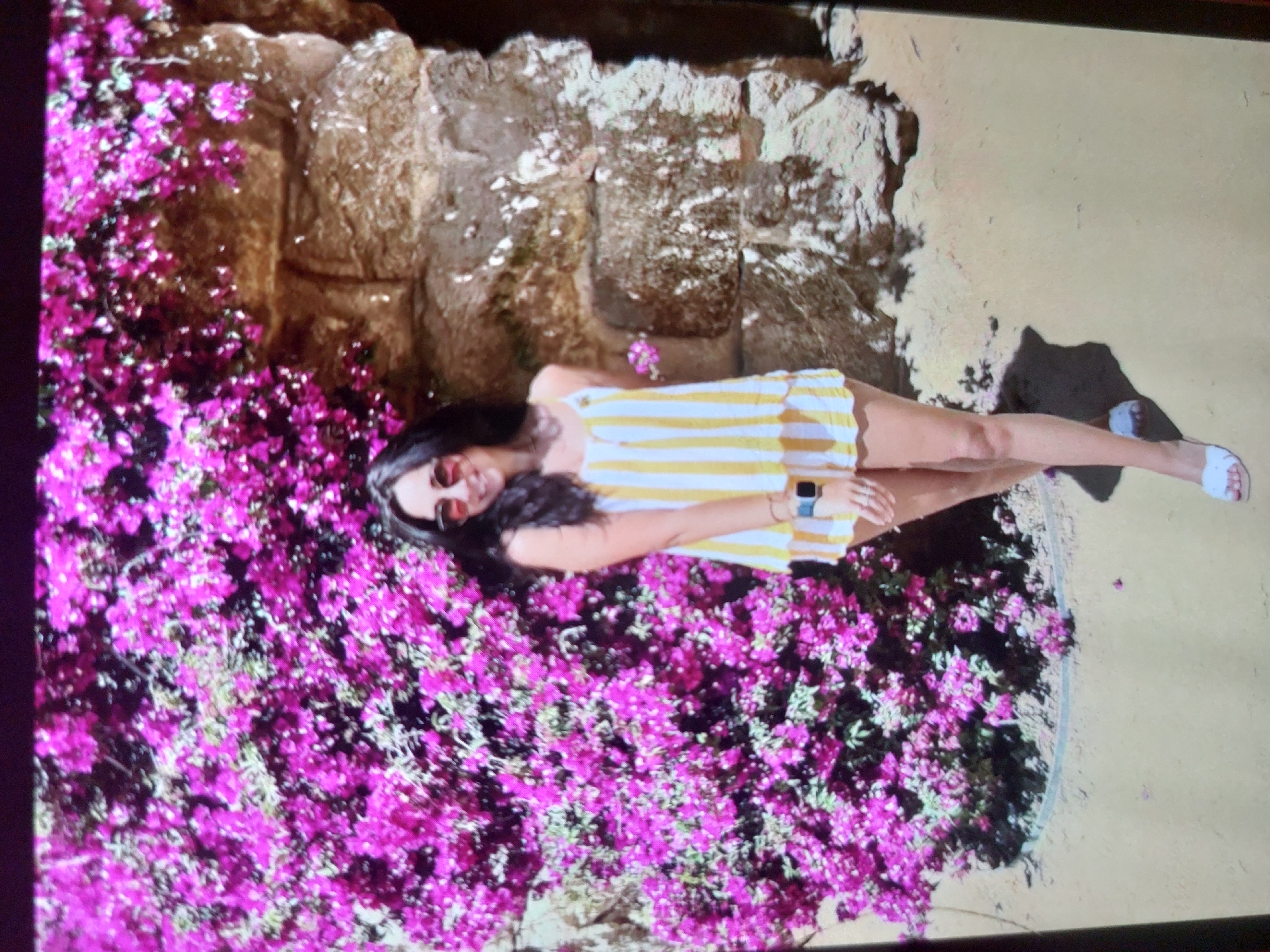 A young woman is captured in a vibrant, sunlit outdoor scene, rotated 90 degrees clockwise. She lies on her back on a light-colored ground, possibly sand or light-colored concrete. She has dark hair, wears sunglasses, and is dressed in a yellow and white striped strapless dress or romper. A watch is visible on her left wrist, and she wears white sandals on her feet.
She is positioned next to a dense, sprawling bush or vine, heavily laden with bright pink/magenta flowers, likely bougainvillea, which occupies a significant portion of the left side of the frame. Behind her head and extending into the background is a rough, textured stone wall or rock formation. The lighting is bright and sunny, casting distinct shadows from the woman and the stone structure onto the ground, indicating it is daytime with clear weather. She appears relaxed and is looking towards the camera with a subtle smile.
The setting, with its abundant bougainvillea and stone features, suggests a warm, possibly coastal environment, consistent with the provided context of Tsarevo, Bulgaria. There is no visible text in the image.