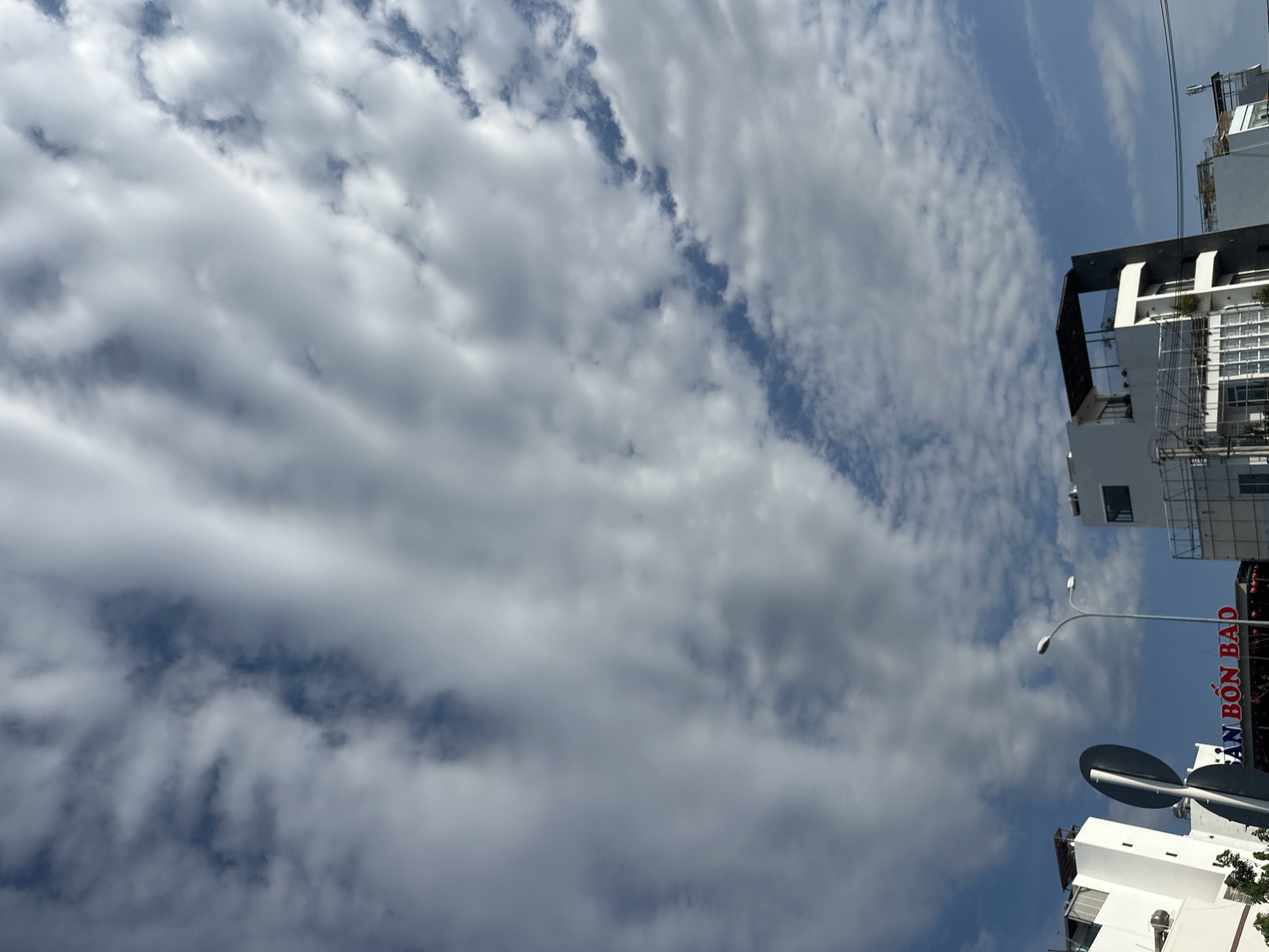 The image shows a view of the sky with altocumulus clouds over Nha Trang Ward, Vietnam. Part of a white building is visible on the right, along with a street light and a traffic sign. Red text, 'AN BON BAO,' is seen on a storefront below the white building. The focus is on the clouds filling most of the frame.