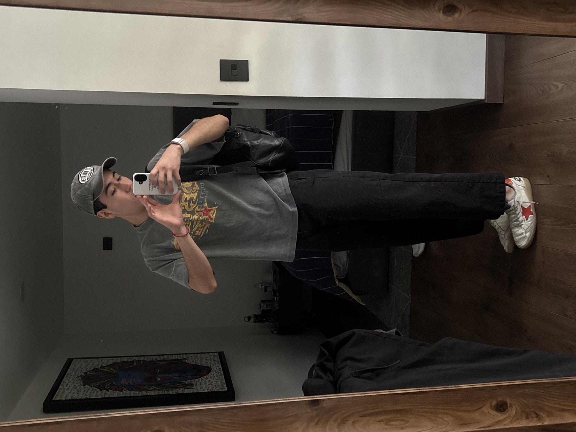 A young man stands in a bedroom, reflected in a full-length mirror. He is wearing a gray baseball cap, a gray t-shirt with a graphic print, black pants, and white sneakers with a red star. He is holding a white smartphone in his right hand, taking a selfie, and his left hand is forming a heart shape. A black backpack is slung over his left shoulder. The bedroom appears to be furnished with a bed with blue and white striped bedding, a nightstand with items on it, and a framed picture on the wall. The floor is made of dark wood. The location is identified as San Bernardino Tlaxcalancingo, Mexico.
