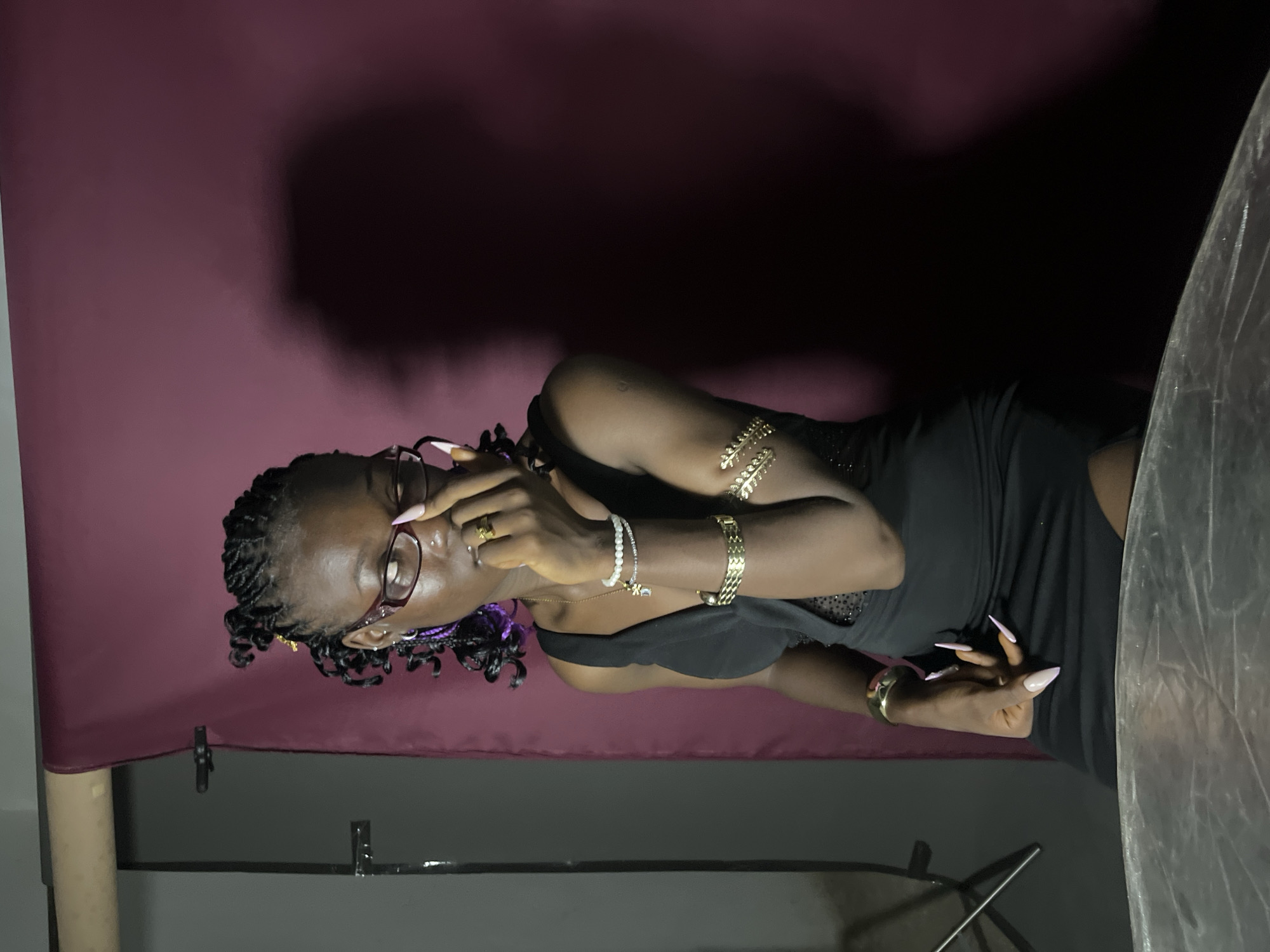 A woman with dark skin and braided hair, adorned with purple extensions and gold accents, is seated in front of a dark red backdrop. She is wearing glasses with dark frames, a black dress with a V-neckline and sheer, embellished inserts, and several pieces of gold jewelry, including bracelets on her upper arms and wrist, and rings on her fingers. Her right hand is raised to her face, obscuring her eye. The lighting casts a shadow of the woman on the backdrop to her right. The setting appears to be a studio with photographic equipment visible on the left side of the frame. The foreground shows a portion of a reflective surface, possibly a table. The overall scene is dimly lit, with the main subject illuminated from the front and slightly to the side. The specific location in Gura Top, Nigeria, cannot be determined from the visual information provided.