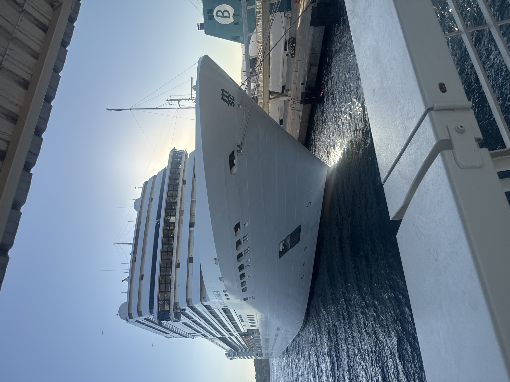 A large, white cruise ship, identified by the name "MSC MUSICA" on its hull, is docked in the port of Ibiza, Spain. The sun is low in the sky, casting a warm light and creating reflections on the water. The ship takes up a significant portion of the frame, with its bow pointed towards the right. In the background, another ship with a green hull and a large white "B" is visible, along with a walkway connecting the ships to the dock. Several mooring lines secure the MSC Musica to the pier. The foreground shows a white railing, suggesting the photo was taken from another vessel or a nearby structure. There are no people clearly visible in the immediate vicinity of the ship, though figures can be seen on the dock in the distance. The water is a deep blue, with gentle ripples. The sky is clear and blue, with a few distant birds. The overall scene depicts a tranquil moment in a bustling port.