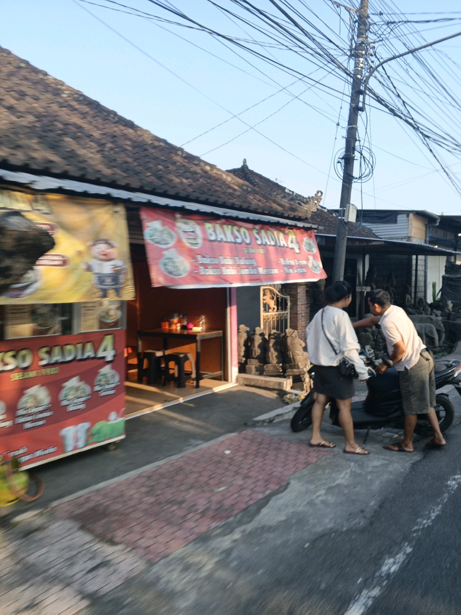 In Sukawati, Indonesia, two individuals are interacting next to a scooter parked on a street. The woman, wearing a white long-sleeved shirt and a black skirt, stands with her back to the camera, facing the man. The man, dressed in a light-colored t-shirt and shorts, is leaning towards the scooter, his right hand extended as if reaching for it or talking to the woman.
The scene is set in front of a row of businesses. To the left, a food stall with red and yellow banners advertises "Bakso Sadia 4." One banner features a cartoon of a plump man in traditional Indonesian attire. Inside the stall, a table and stools are visible, with condiments on the table. To the right of the food stall, a row of stone statues are displayed, including what appear to be elephants and other figures. Further down the street, more businesses with dark facades are visible, one with a prominent utility pole densely packed with electrical wires. The street itself is paved with asphalt and features a section of red brick paving. The overall lighting suggests it is late afternoon or early morning.
