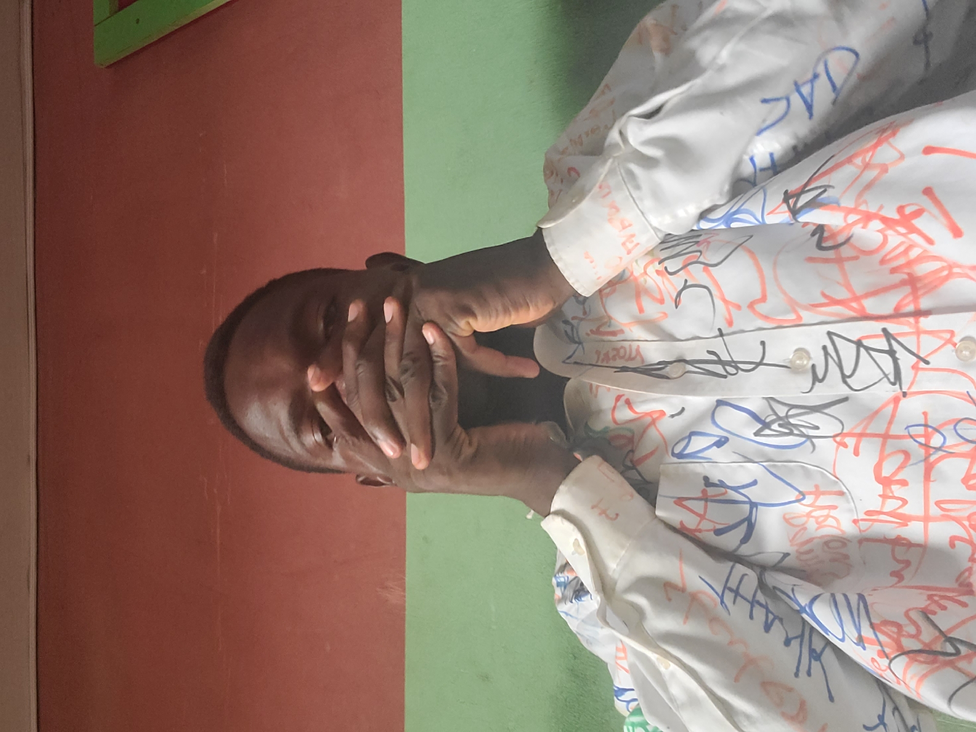 A close-up shot shows a person of African descent covering their mouth and nose with their hands, obscuring their face. They are wearing a white collared shirt adorned with colorful scribbled markings in red, blue, and black. The background consists of a wall divided horizontally into two sections: the upper portion is a reddish-brown, and the lower portion is light green. A sliver of a green object is visible on the right side of the frame, suggesting an outdoor or semi-outdoor environment. The lighting appears to be natural, possibly from a window or open space. No specific location in Gura Top, Nigeria, can be identified from the image.