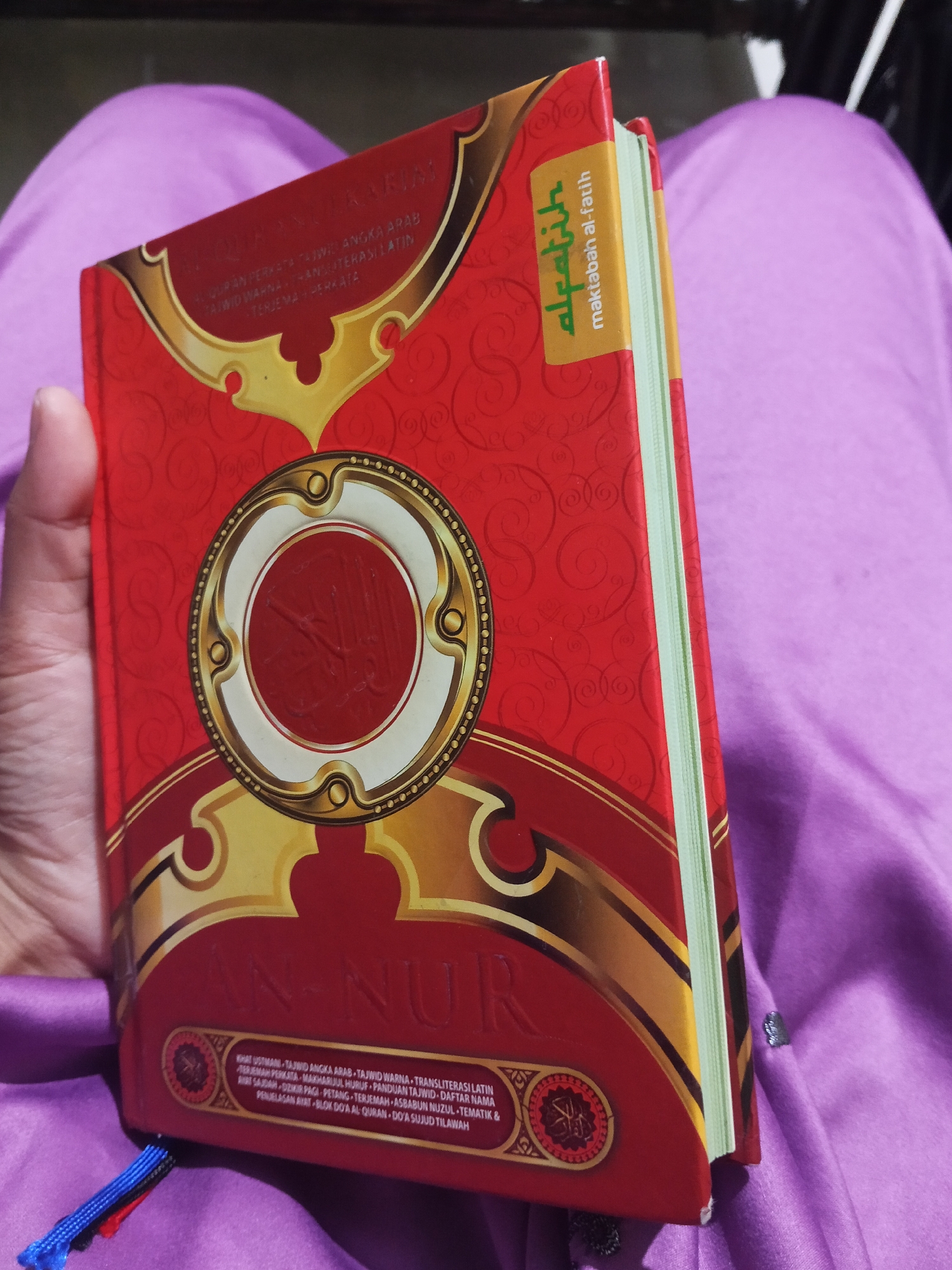 A person's hand is holding a red book, which appears to be a copy of the Quran. The book has ornate gold and white designs on its cover and spine. The text on the cover includes Arabic calligraphy and what looks like Indonesian or Malay. A label on the spine reads "al-Fatih" in Arabic and "maktabah al-Fatih" in Latin script. The book is resting on a purple fabric, possibly a hijab or prayer mat. The background is dark and out of focus. The scene is indoors. There are no identifying landmarks or elements that would allow for precise location within Lestari Indah, Indonesia.