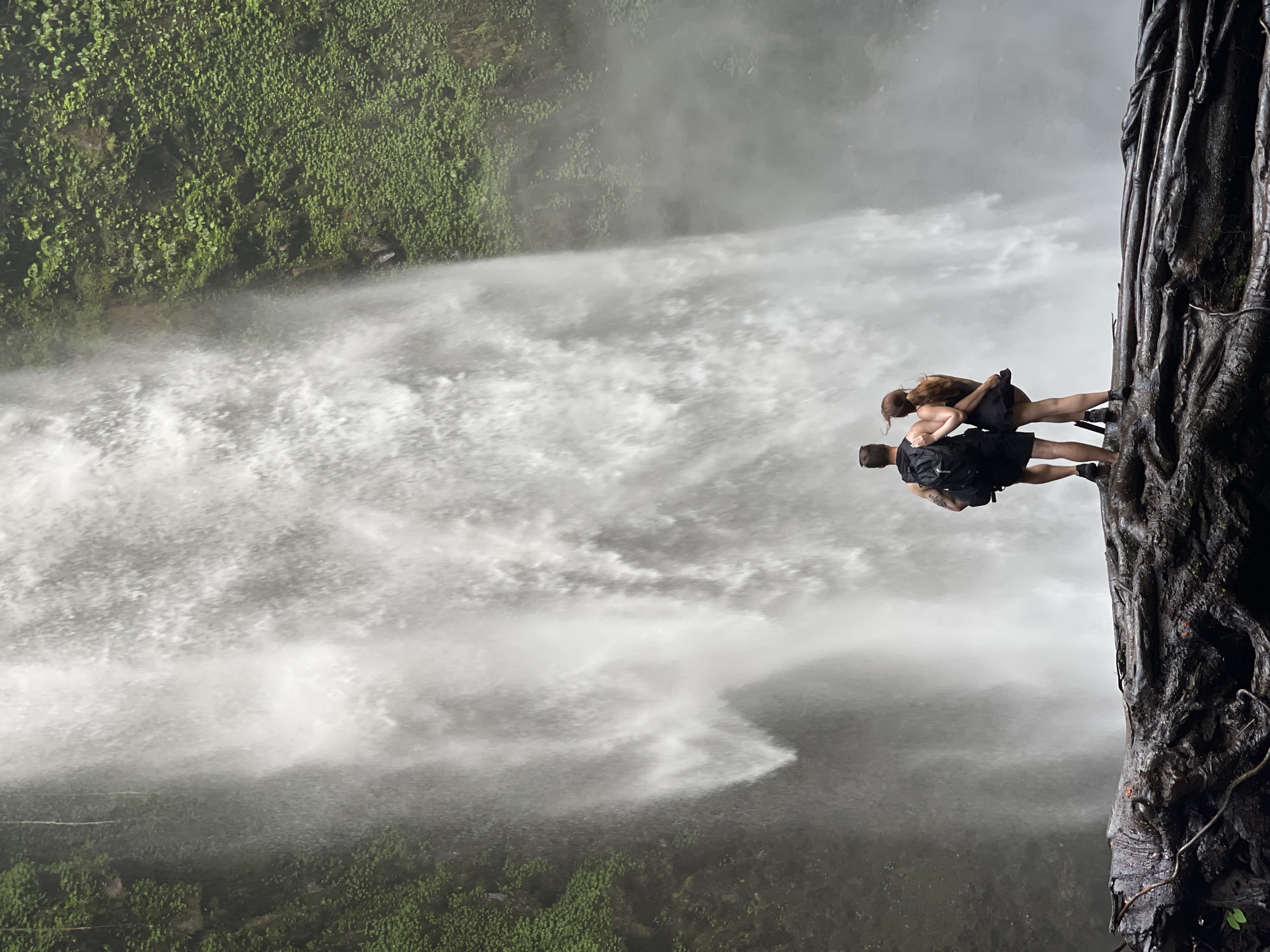 In Petang, Indonesia, a couple stands on a fallen, moss-covered tree trunk, facing away from the camera. They are positioned in the foreground with a towering waterfall as their backdrop. The man, on the left, wears a black backpack and shorts. The woman, on the right, is dressed in a black dress. The waterfall cascades down a lush, green cliff face, creating a misty atmosphere. The scene captures a moment of quiet appreciation for the natural beauty of the Indonesian landscape.