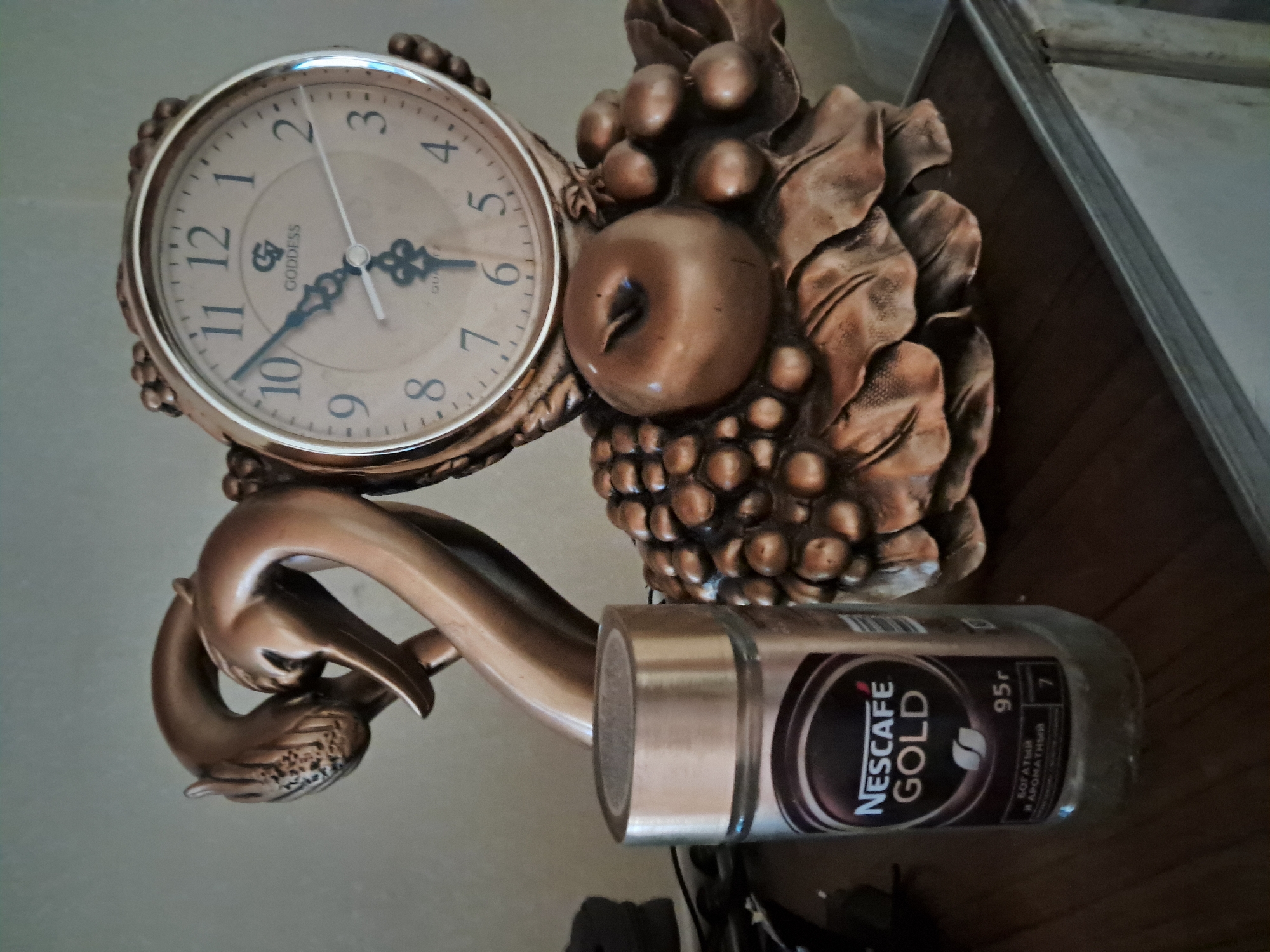 A close-up, eye-level, indoor shot features a decorative table clock with a bronze finish. The clock face is round with Arabic numerals and has a "Goddess" logo. The clock is adorned with sculpted grapes and an apple. To the left of the clock, a jar of Nescafé Gold instant coffee is visible, with Russian text on the label. The items are placed on a dark wooden surface. The background is a plain, light-colored wall. The scene appears to be set within a home, with no indication of being in Bagish, Uzbekistan.