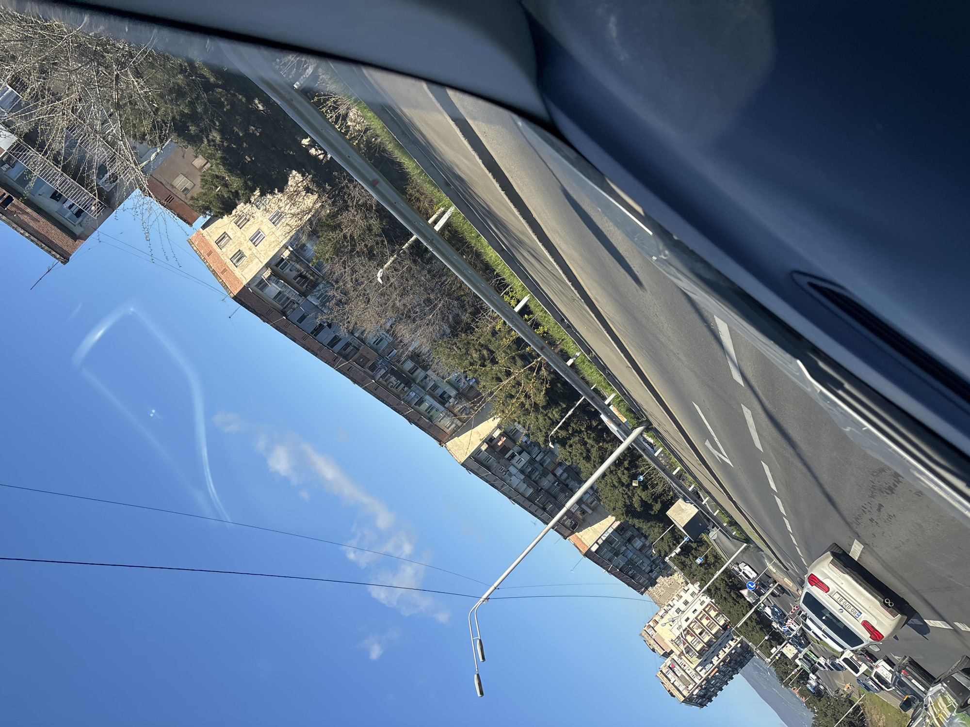 The image, taken from inside a vehicle, shows an urban street scene in Tbilisi, Georgia. The road, marked with white lines, is filled with vehicles, including cars and buses. Multi-story residential buildings line the street. The sky is clear and blue. A street light is visible.