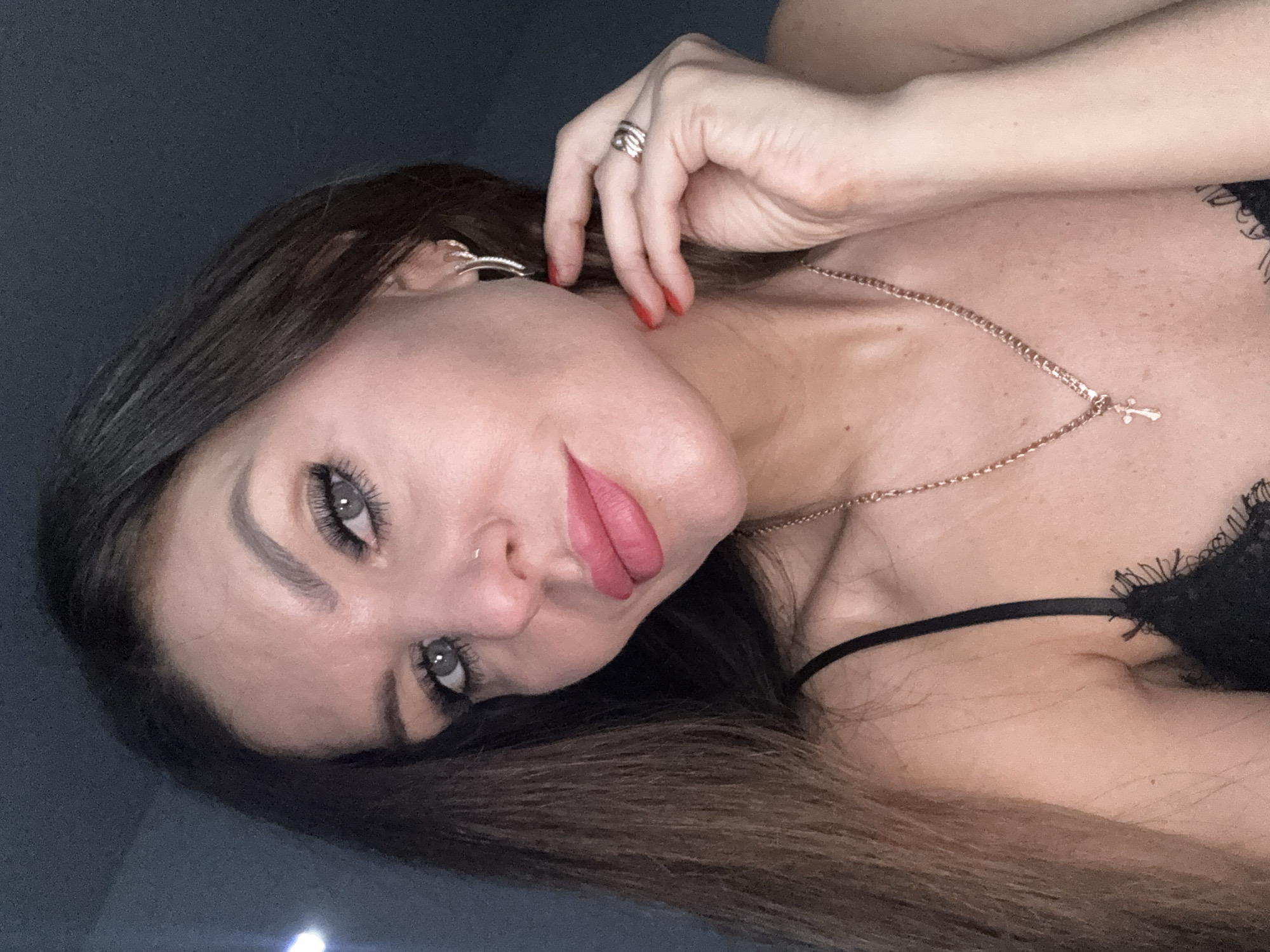 A woman is posing for a selfie. She has long brown hair, light skin, and gray eyes. She is wearing a gold necklace with a cross pendant, a ring on her finger, and what appears to be a black lace-trimmed garment. She is positioned against a dark background. The location is unspecified and unrelated to Chaikovsky, Russia.