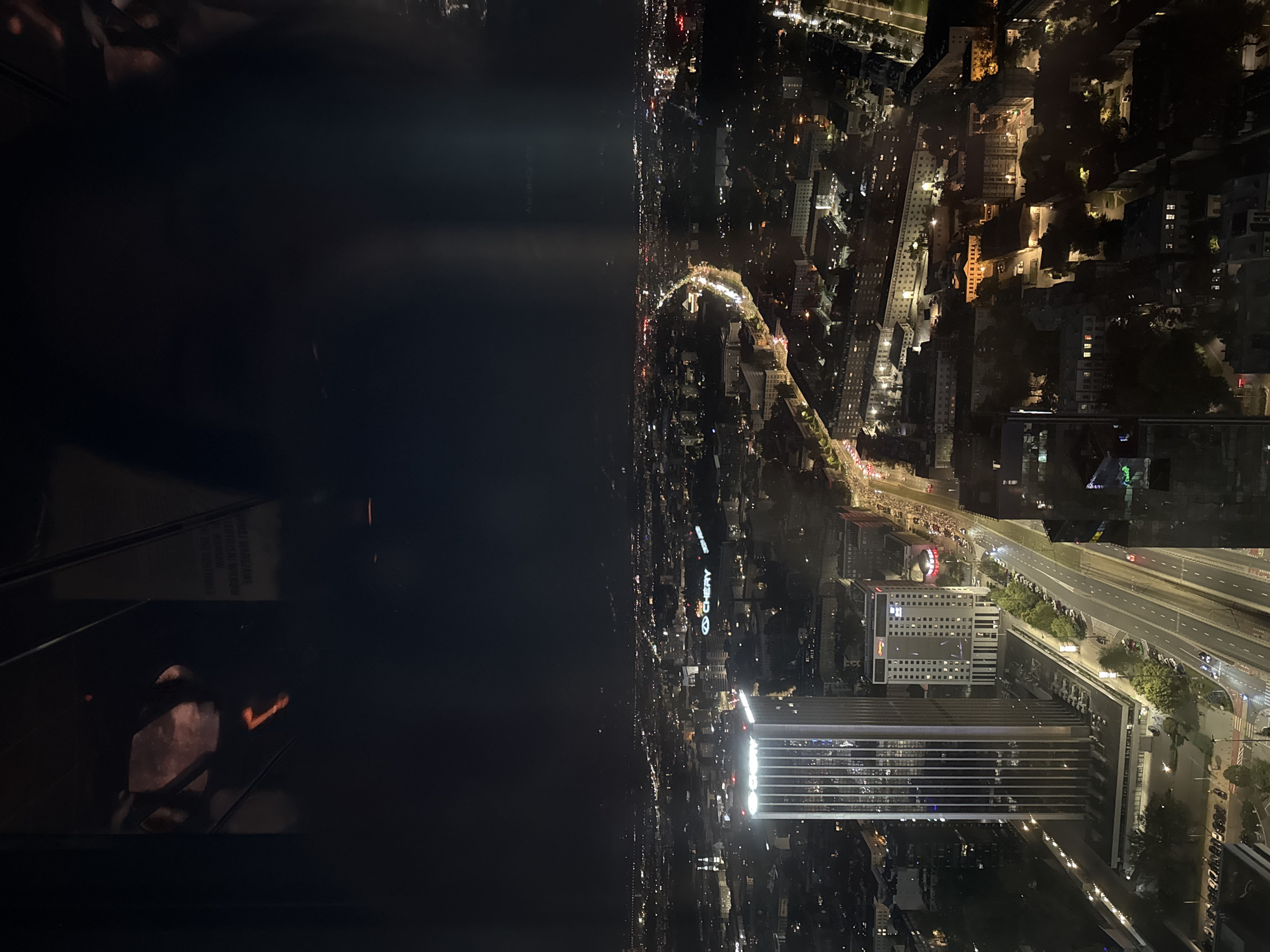 The image captures an elevated night-time view of Warsaw, Poland. The foreground features the brightly lit facade of the Skylight building, a prominent skyscraper. Adjacent to it is the Hampton by Hilton Warsaw City Centre hotel. A wide, multi-lane road runs in front of these buildings, with cars visible on the road. The cityscape extends into the distance, characterized by a dense arrangement of buildings with illuminated windows, creating a network of city lights. A major road, likely Aleje Jerozolimskie or a similar arterial route, curves through the urban landscape, with traffic illuminated along its length. In the upper portion of the image, the sky is dark, with faint vertical light beams suggesting artificial lighting or atmospheric effects. Reflections are present in the foreground, hinting at the viewpoint being from inside a building or vehicle with windows. The overall scene depicts a bustling urban environment at night, showcasing the architecture and infrastructure of Warsaw.