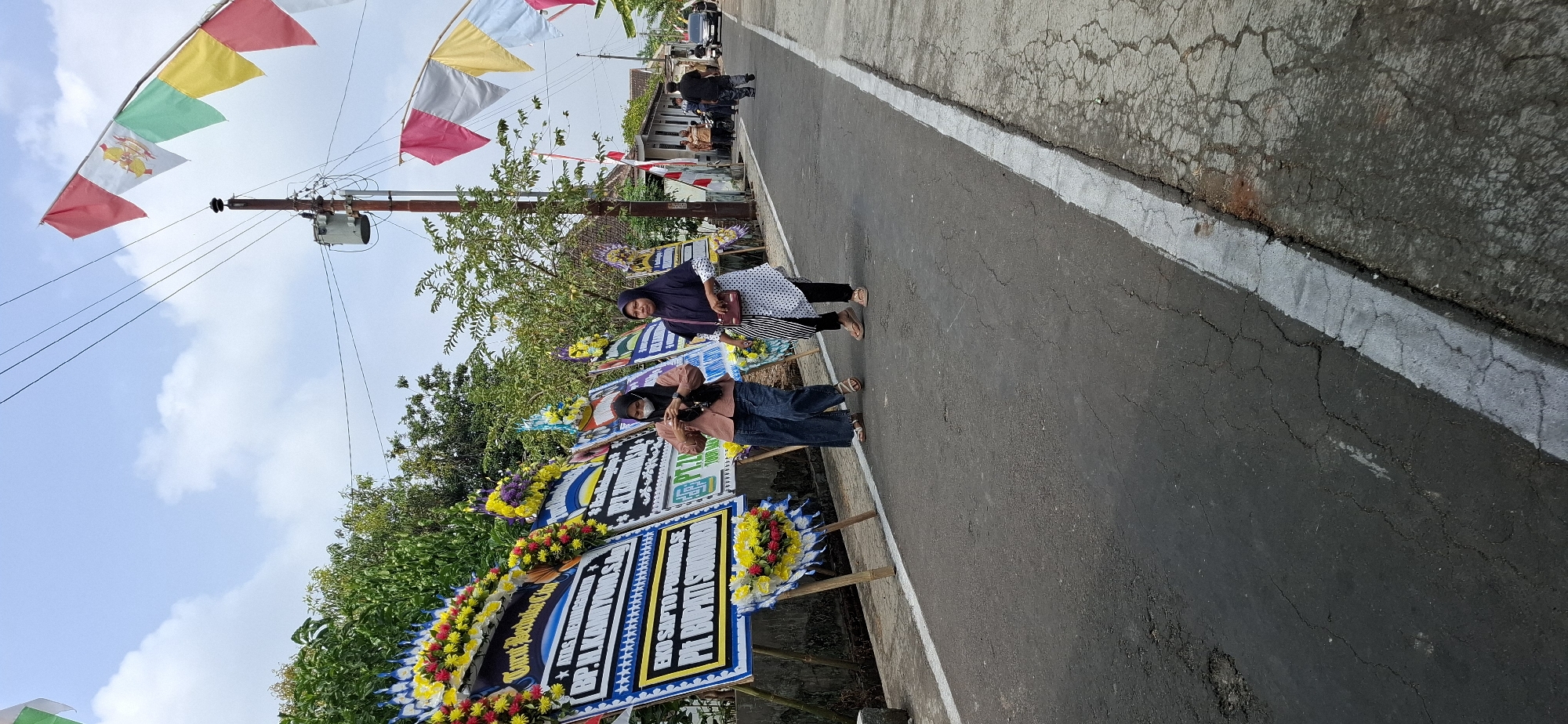 In Gentan, Indonesia, a road is lined with numerous elaborate floral arrangements, commonly known as "papan bunga" or flower boards, typically used for condolences or congratulations. Two women are walking along the road, away from the camera, with the flower boards on their left. The woman in the foreground, on the right, wears a navy blue hijab and a white polka dot top over black pants, carrying a small maroon purse. The woman to her left wears a light brown hijab and a matching shirt with dark blue jeans.

In the background, more flower boards are visible, as well as a residential street with houses. A utility pole with a transformer stands prominently near the center of the image, adorned with a string of colorful triangular flags. Several people are walking further down the street, and a car and a motorcycle are also present. The environment appears to be a sunny day with a partly cloudy sky, and the road shows signs of age with visible cracks and wear.