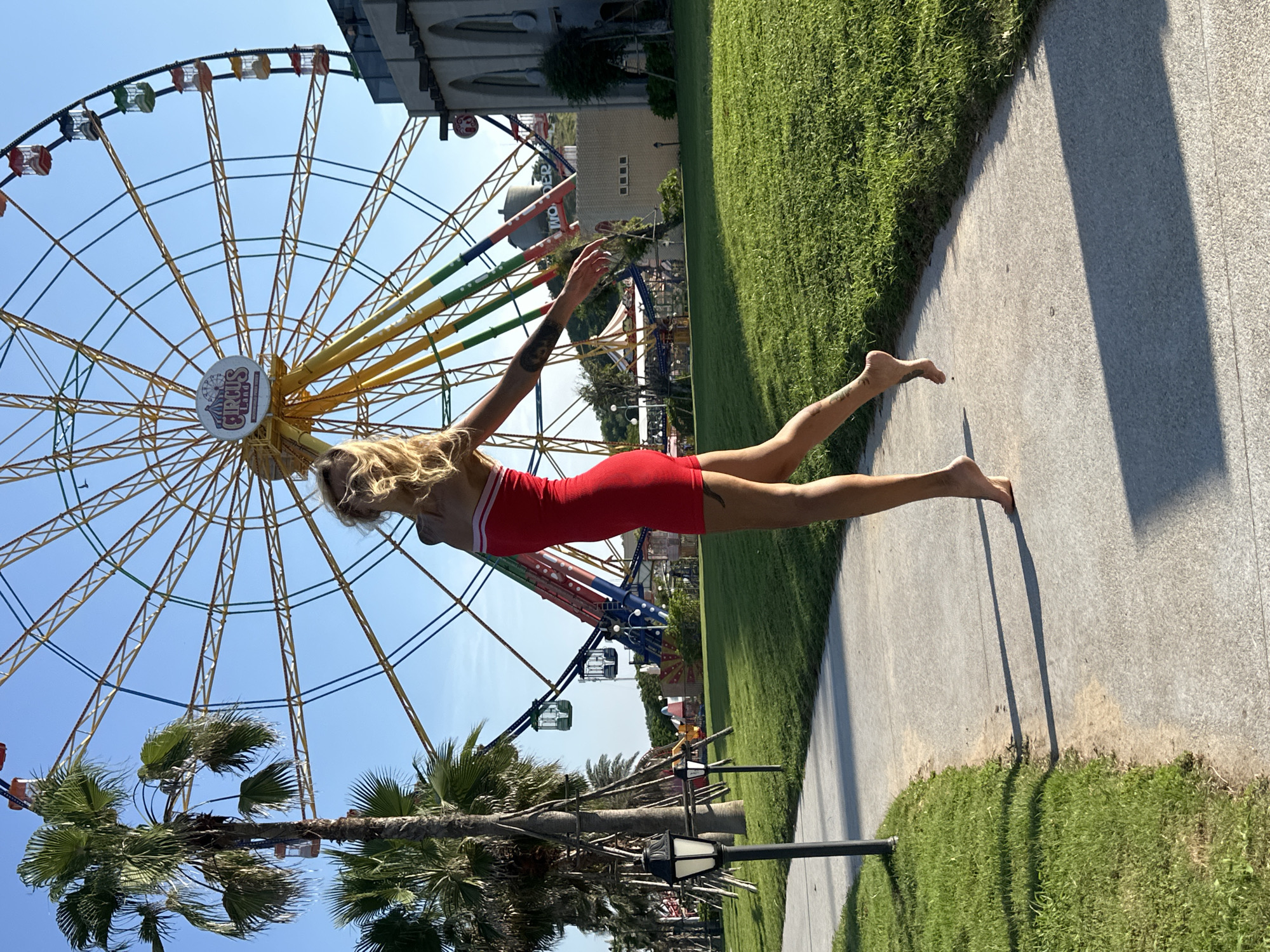 In the city of Tien Thanh Ward, Vietnam, on a sunny day, a young woman with blonde hair, wearing a red strapless dress, poses barefoot on a concrete path. She is captured mid-stride, with her left leg extended and her right leg bent, appearing to jump or dance. The path runs alongside a well-maintained green lawn. In the background, a large, colorful Ferris wheel with various cabins is prominently visible, along with palm trees and buildings. The Ferris wheel has a sign that reads "CIRCUS LAND." The scene suggests an amusement park or recreational area.