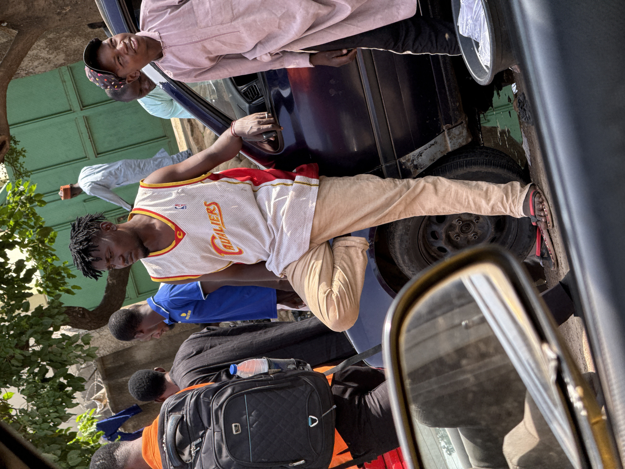 The image, taken from inside a vehicle and originally rotated 90 degrees counter-clockwise, depicts a vibrant street scene in what appears to be Abuja, Nigeria, under bright daylight. It captures a gathering of several young Black men around a parked dark-colored car, suggesting a casual social interaction or a moment of waiting.
In the central foreground, a young man with dreadlocks wears a white basketball jersey with "CAVALIERS" visible on the chest in red and yellow, light beige pants, and red flip-flops. He is leaning against the passenger side of a dark blue or black sedan, with his right hand resting near the door handle.
Leaning out of the car's passenger window above him, another young man in a light pink long-sleeved shirt and a colorful patterned baseball cap looks upwards with a slight smile. A third man in a light blue shirt is partially visible behind him.
Further to the left, several other men are present. One wears a blue polo shirt, while another in a black t-shirt carries a black quilted backpack topped with a blue-capped water bottle. An additional man in an orange t-shirt with a backpack is also visible. In the background, a man in a light-colored traditional garment and an orange head covering walks away.
The parked car shows some visible wear near its rear wheel. To the left, there is lush green foliage from a tree, and in the background, a distinctive green corrugated metal gate or wall. The scene is brightly lit by the sun, indicating clear weather. The side-view mirror of the car from which the photo was taken is partially visible in the bottom right, obscuring a small portion of the scene. The overall mood appears relaxed and social.