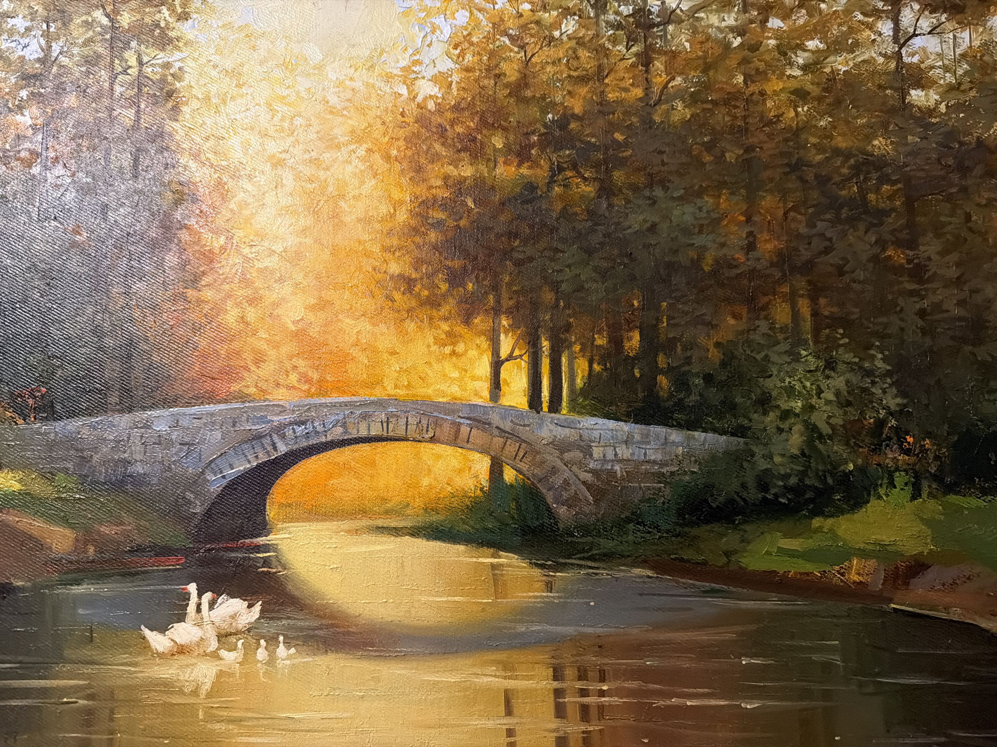 The image is an oil painting of a picturesque landscape. A stone bridge arches over a calm body of water. Sunlight streams through a dense canopy of trees, casting a warm, golden glow on the scene and illuminating the water. On the left side of the water, a family of swans, including two adults and several cygnets, are swimming. The environment appears to be a park or natural setting. The specific location in the city of Bakikhanov, Azerbaijan, is not discernible from the visual information provided in the painting.