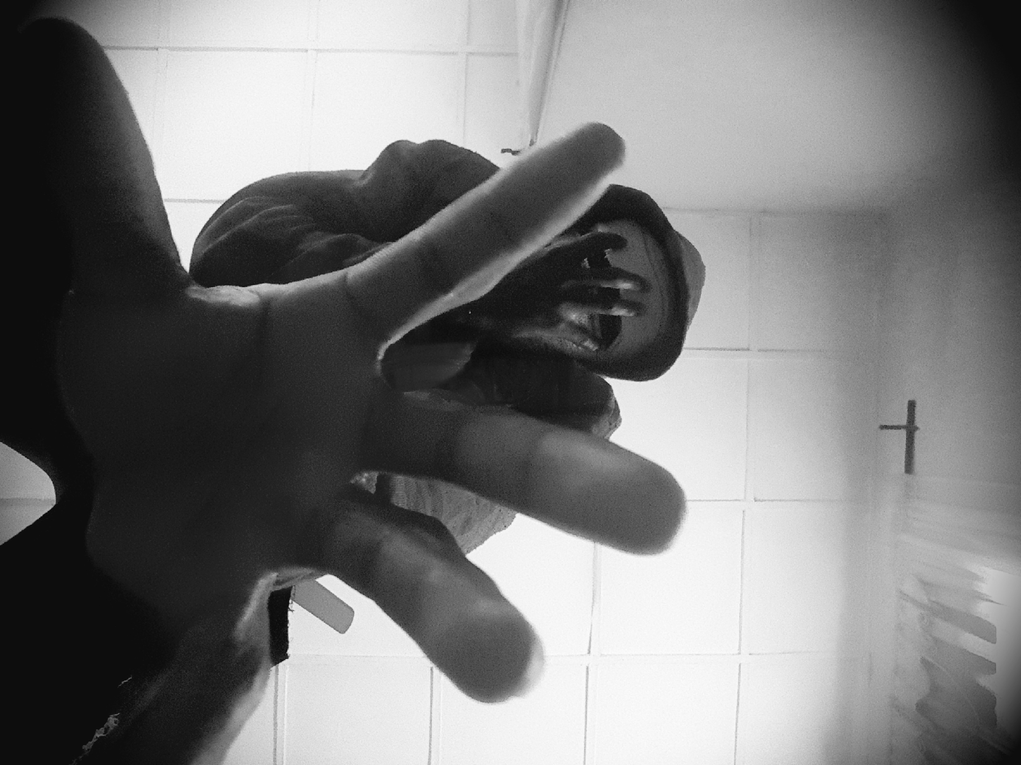 A monochrome, low-angle photograph, captured in Narayi, Nigeria, depicts a single individual seen from a low perspective. The subject, wearing a dark hooded garment that heavily shades their face, extends their right hand prominently forward and downward, with fingers splayed in a distinct reaching or grasping gesture towards the camera's position. Their left hand is partially visible further back, near the obscured facial area. The background consists of light-colored, square-tiled walls or a ceiling, forming a grid pattern. A small, dark, upright cross is affixed to a tiled wall in the lower-central portion of the frame. The scene is illuminated by ambient light, creating strong contrasts that emphasize the subject's silhouette against the brighter background. The perspective exhibits noticeable wide-angle distortion.