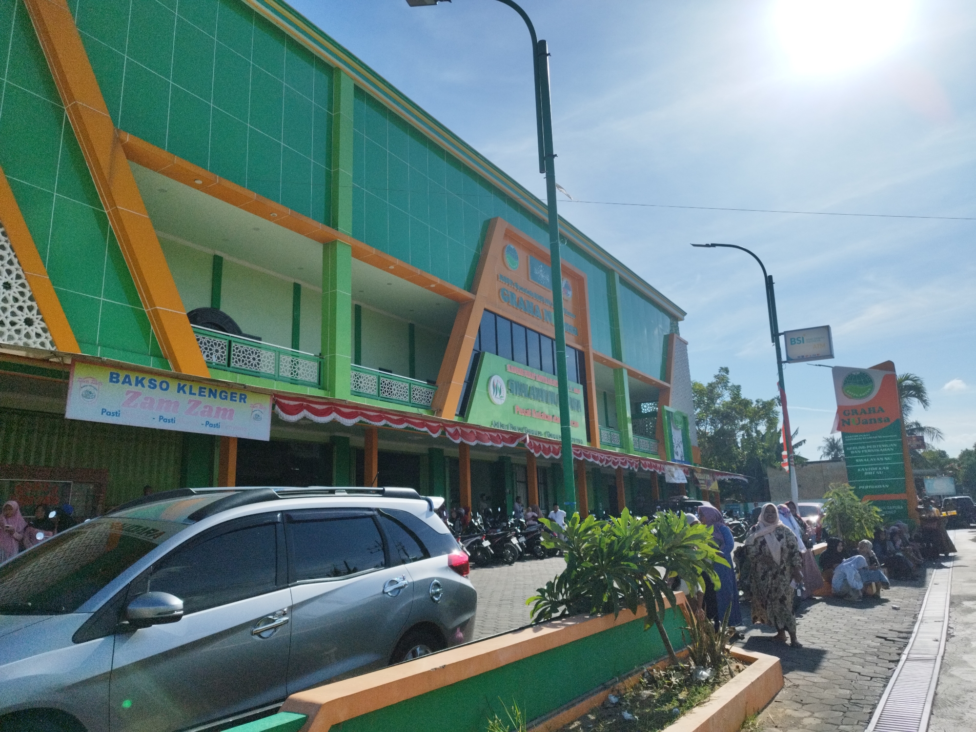 This is a daytime, eye-level, outdoor, medium-shot image taken in front of a multi-story building with a green and orange facade in Mandala, Indonesia. The building is modern with a tiled exterior and prominent orange diagonal beams. Various signs are visible on the building, including one that reads "GRAHA N JANSA" and another advertising "BAKSO KLENGER Zam Zam". A silver car is parked in the foreground to the left, with tinted windows. To the right, several people are gathered near the building's entrance and on the sidewalk, some sitting and some standing. A variety of motorcycles are parked in front of the building. The sky is bright blue with the sun prominently displayed, suggesting a sunny day. A street lamp and electrical wires are also visible. The ground is paved with interlocking bricks. Lush green plants are growing in planters along the sidewalk.