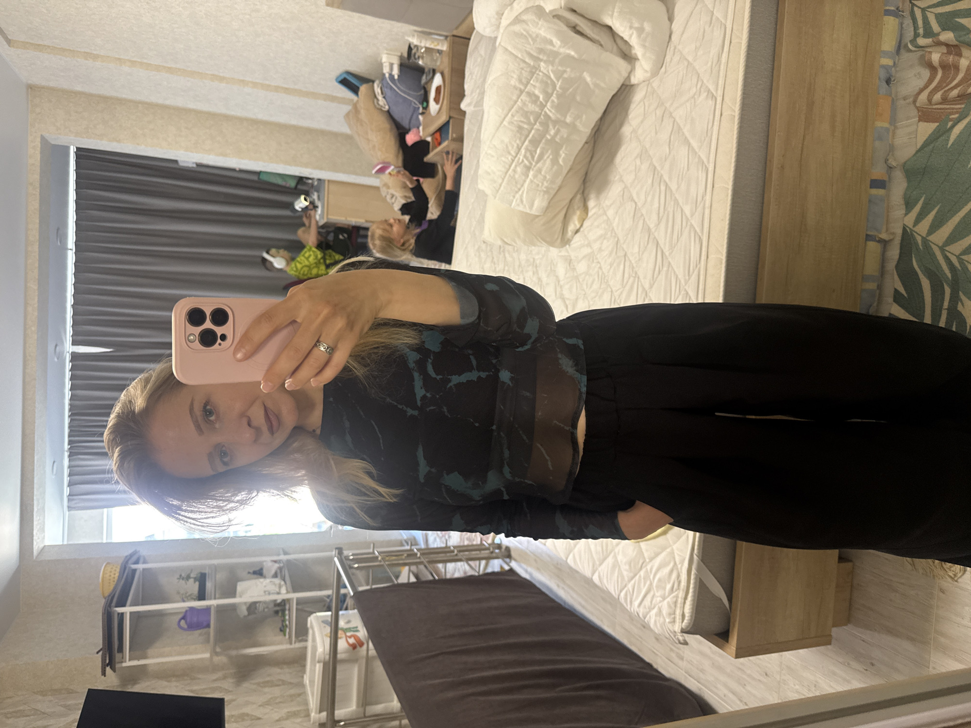 A mirror selfie of a woman with blonde hair, wearing a black top and pants, is seen in an indoor setting. The reflection reveals a room with a bed, a shelf with various items, and a window with blinds. The woman is holding a pink smartphone to take the selfie. Other people and objects can be seen in the mirror's reflection. The location is unspecified.