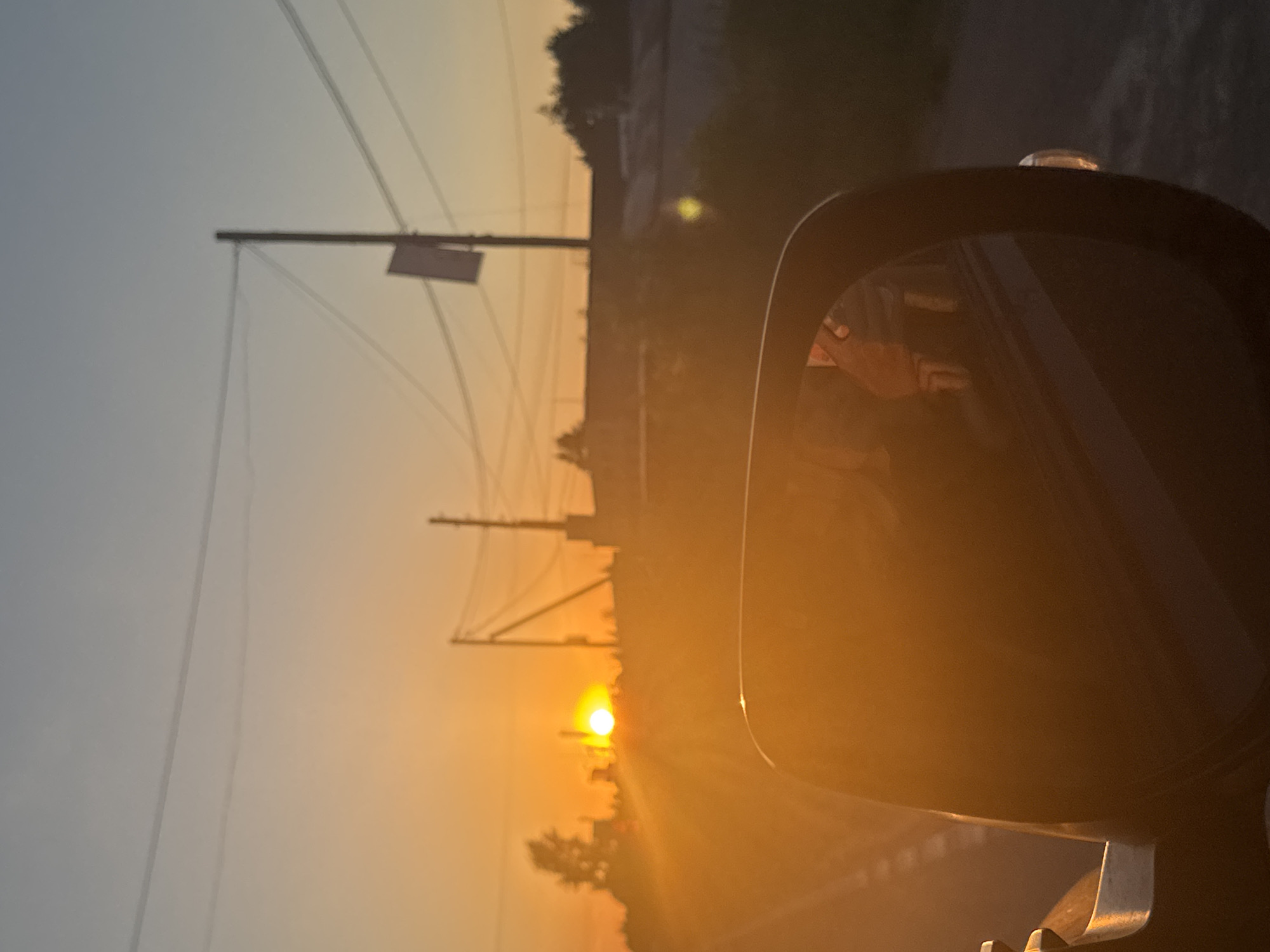 The image captures a view of a sunset, likely taken from a vehicle in Chaikovsky, Russia. The sun is a bright, orange orb low on the horizon, casting long rays of light across the scene. In the foreground, a portion of a car's side-view mirror is visible, reflecting a blurred image of what appears to be the interior of the car and possibly a passenger. Beyond the mirror, a road stretches into the distance, bordered by a faint white line. Silhouetted against the sunset are trees and buildings, with utility poles and power lines crisscrossing the sky. The overall atmosphere is warm and hazy due to the low angle of the sun.
