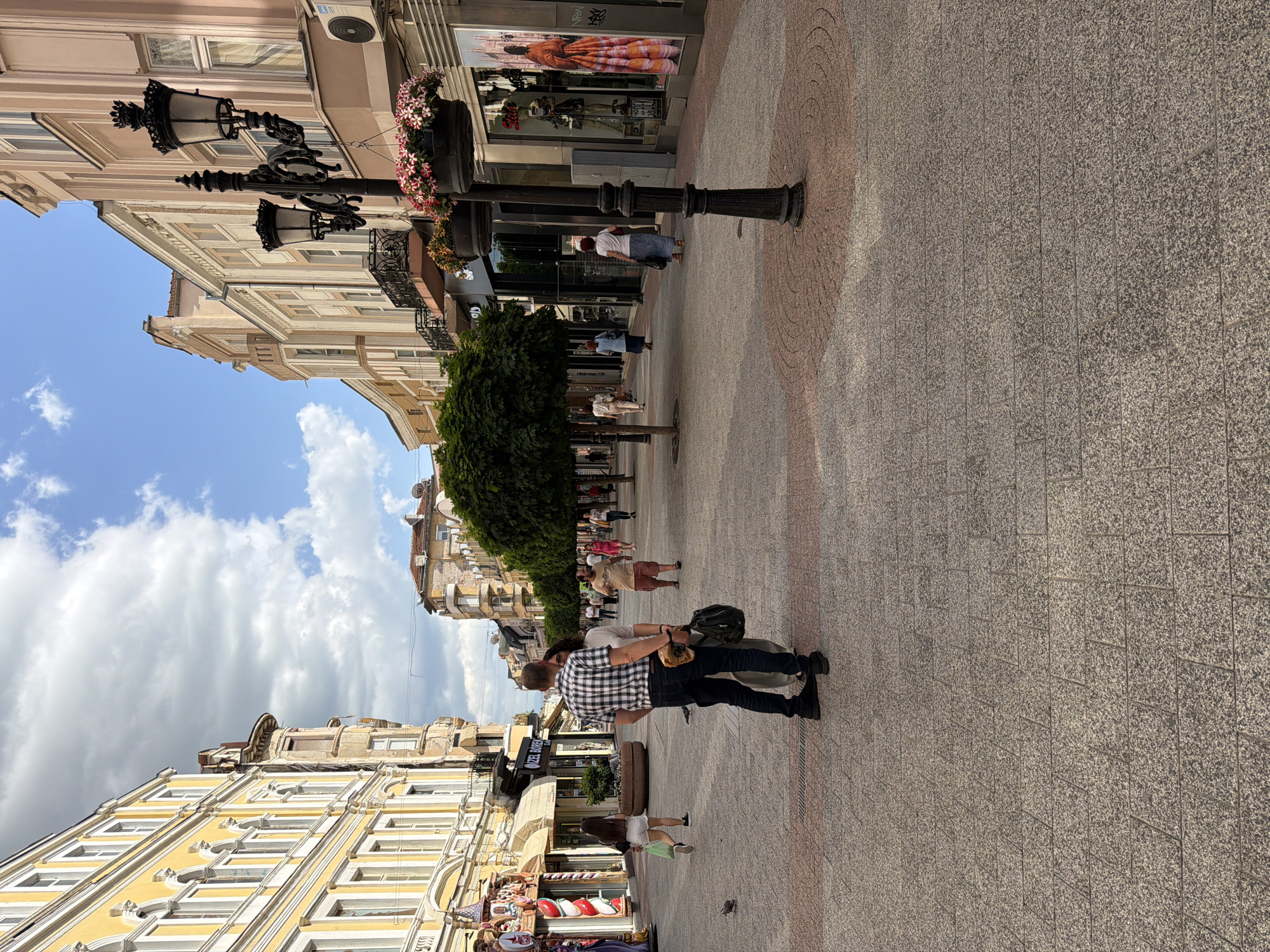 The image captures a street scene in Plovdiv, Bulgaria. Pedestrians are walking on a paved street lined with buildings. The architecture is primarily historical, with facades of beige and yellow buildings and some elaborate window designs. Some lampposts with flower arrangements are interspersed along the walkway. The sky is blue with scattered clouds. A couple is walking along the street. Shops are visible in the background with advertisements.