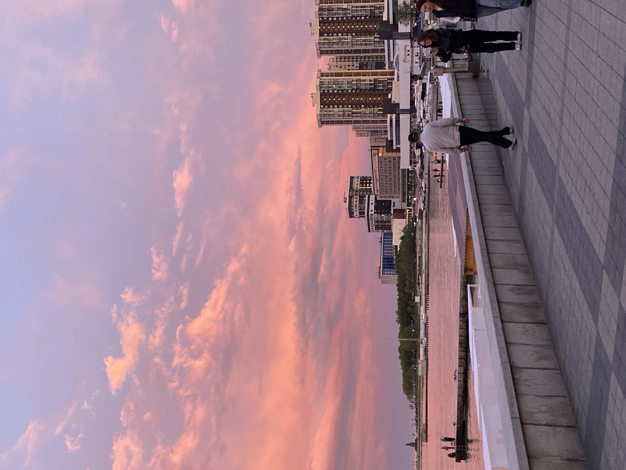 This image depicts a scenic waterfront promenade at either sunset or sunrise, likely in Kabardinka, Russia. The sky dominates the upper portion, showcasing a breathtaking display of pink, orange, and purple hues with scattered clouds, indicating the early or late hours of the day.
Below the vibrant sky, a calm body of water, possibly a sea or a large river, mirrors the intense colors, appearing in a reddish-pink tone. Across the water, a modern cityscape is visible, characterized by multiple high-rise buildings with numerous windows, some of which are illuminated. A line of green trees separates the buildings from the water's edge.
In the foreground, a wide promenade paved with alternating light and dark grey square tiles stretches horizontally. A white railing separates the walkway from the water. Several individuals are present on the promenade. Closest to the viewer, a person wearing a light top and dark pants stands with their back to the camera, seemingly admiring the view. Further back, two more people are visible, one in a dark jacket and face mask, and another partially seen beside them, suggesting they are strolling or observing the surroundings.
The scene conveys a peaceful and picturesque atmosphere, emphasizing the beauty of the natural light reflecting off the clouds and water, contrasted with the urban backdrop. No identifiable text is clearly legible in the image.