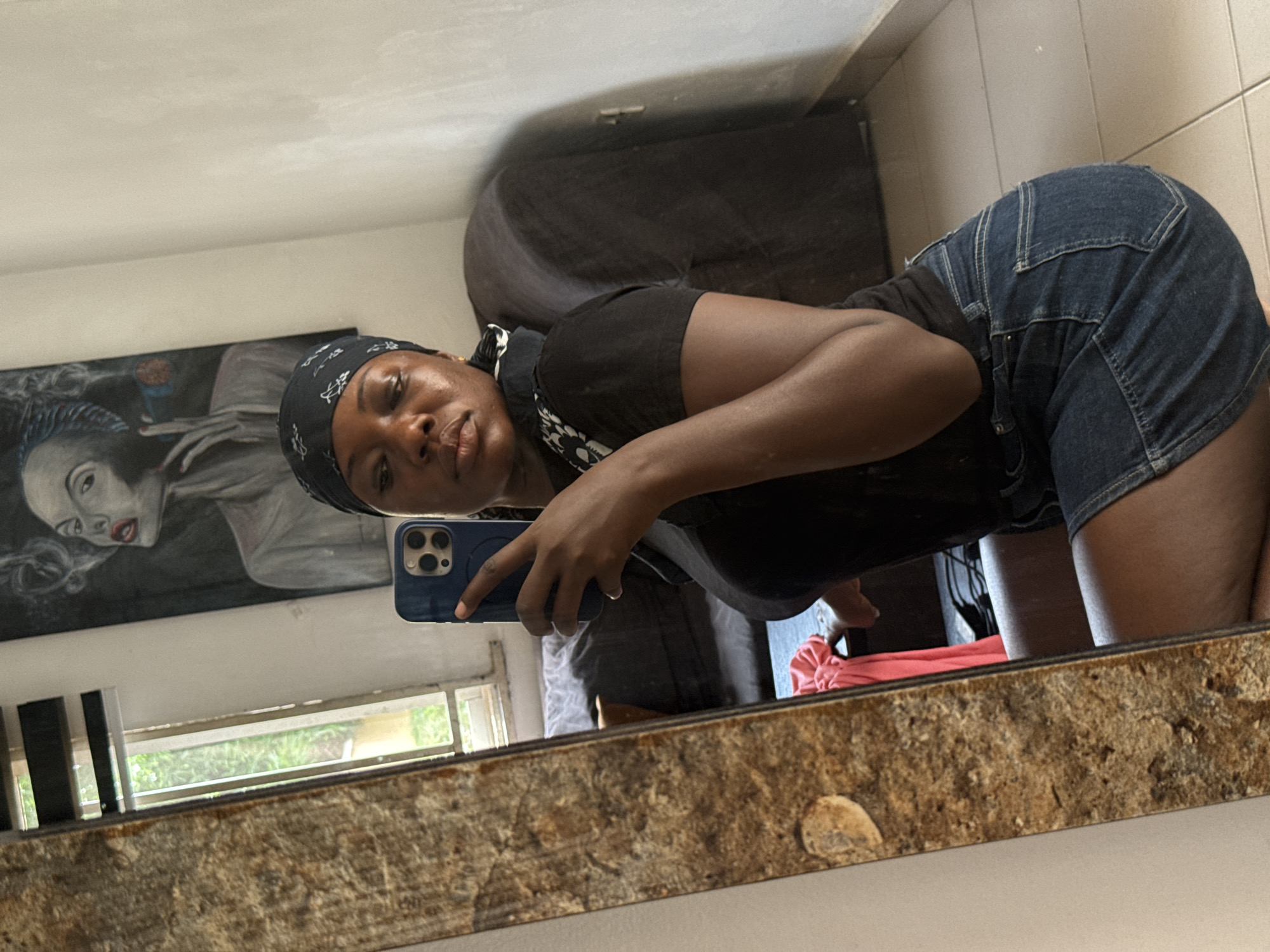 A woman takes a mirror selfie in what appears to be an indoor setting, possibly a bathroom or bedroom. The woman is light-skinned and dressed in a dark t-shirt and denim shorts. She is wearing a headscarf. She is bending at the waist with her buttocks protruding towards the mirror. She holds a blue smartphone, taking the picture. The reflection in the mirror reveals part of the room, including a bed, a framed portrait of a woman, and a window with green foliage outside. The foreground shows the frame of the mirror, which is a textured brown material with spots or patches. The location is not identifiable as Abuja, Nigeria, from the information available in the image.