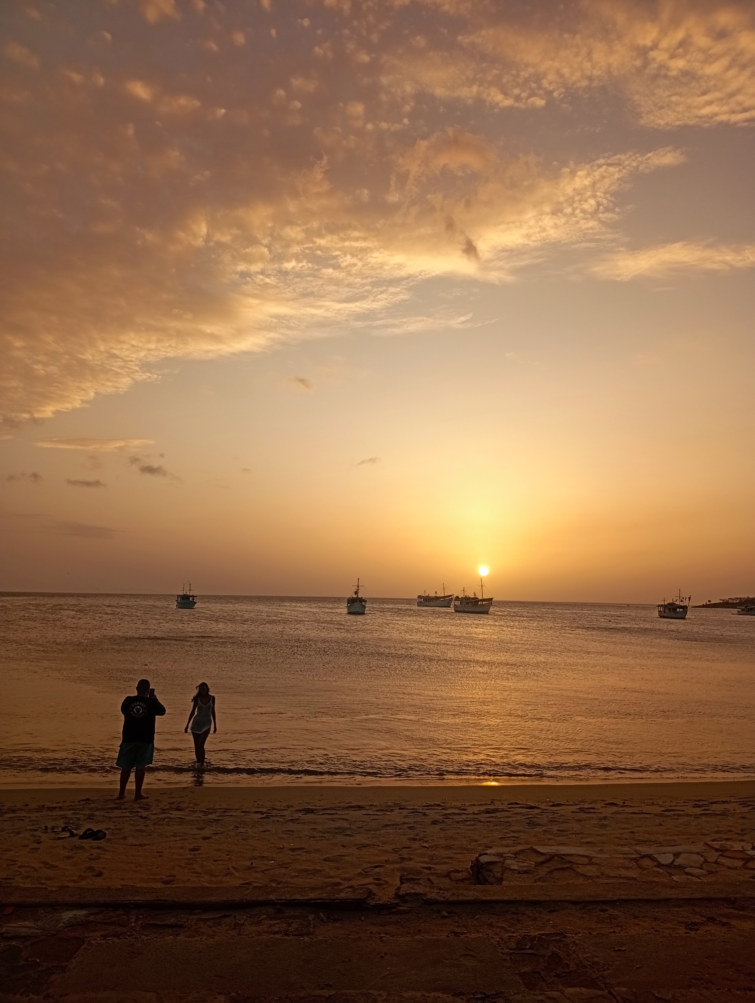 The image presents a picturesque sunset scene over the sea in Juan Griego, Venezuela. The sky is awash in warm hues of orange, gold, and soft pink, with scattered clouds illuminated by the setting sun, which appears as a radiant disk low on the horizon. The sunlight creates a shimmering path across the calm water, extending towards the viewer.
Several boats, appearing to be fishing vessels or small commercial boats, are visible on the water, silhouetted against the bright horizon and the evening sky. They are spread across the mid-ground, some closer to the center and others further towards the left and right edges.
In the foreground, a sandy beach occupies the lower portion of the frame, with gentle waves breaking at the shoreline. Two individuals are present on the beach, silhouetted against the brilliant light. On the left, a person, possibly male, is standing, wearing shorts and a t-shirt with some partially legible text that appears to begin with "SOUND" or "FOUND." This person is holding a device, likely a phone or camera, as if taking a picture. To their right, another person, seemingly female and wearing a light-colored dress or swimsuit, stands in the shallow water, facing away from the camera and looking out towards the sea and the sunset. Two flip-flops are visible on the sand near the person taking the photo.
The scene captures a tranquil moment at dusk, with the two figures enjoying or documenting the natural beauty of the sunset over the serene ocean.