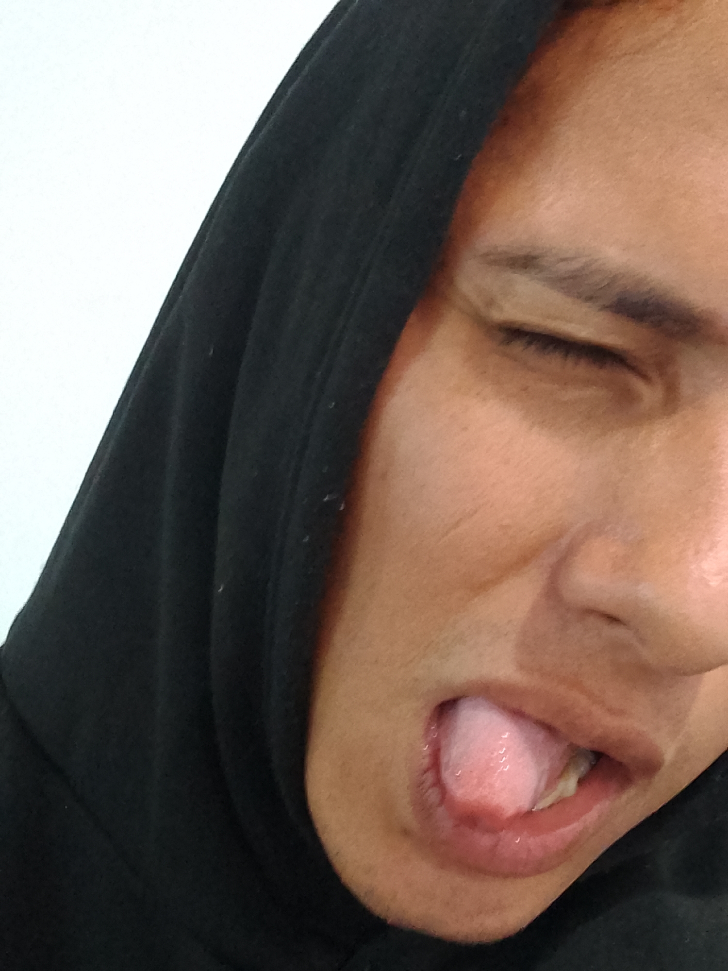 The image presents a close-up, cropped view of an individual's face, taken in a domestic or studio setting within the city of Porlamar, Venezuela. The person, appearing to be an adult with light brown skin, is wearing a black hooded garment, with the hood pulled up and framing the left side of the face. The right eye is closed, consistent with a squint or wink. The mouth is open, revealing a pink tongue partially extended and slightly curled, with some upper and lower teeth visible behind it. The immediate background is a plain, untextured white wall or surface. Lighting is soft and even, highlighting the facial features without harsh shadows. No other objects or individuals are present in the frame.