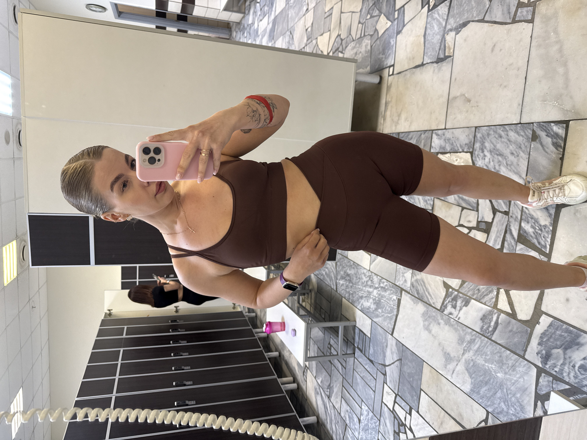 A woman is taking a selfie in what appears to be a locker room. She is wearing a brown athletic top and shorts set, and white sneakers. She is holding a pink phone. There is a person in the background facing away from the camera. Lockers are visible in the background. The floor is marble. The location is identified as Chaikovsky, Russia.