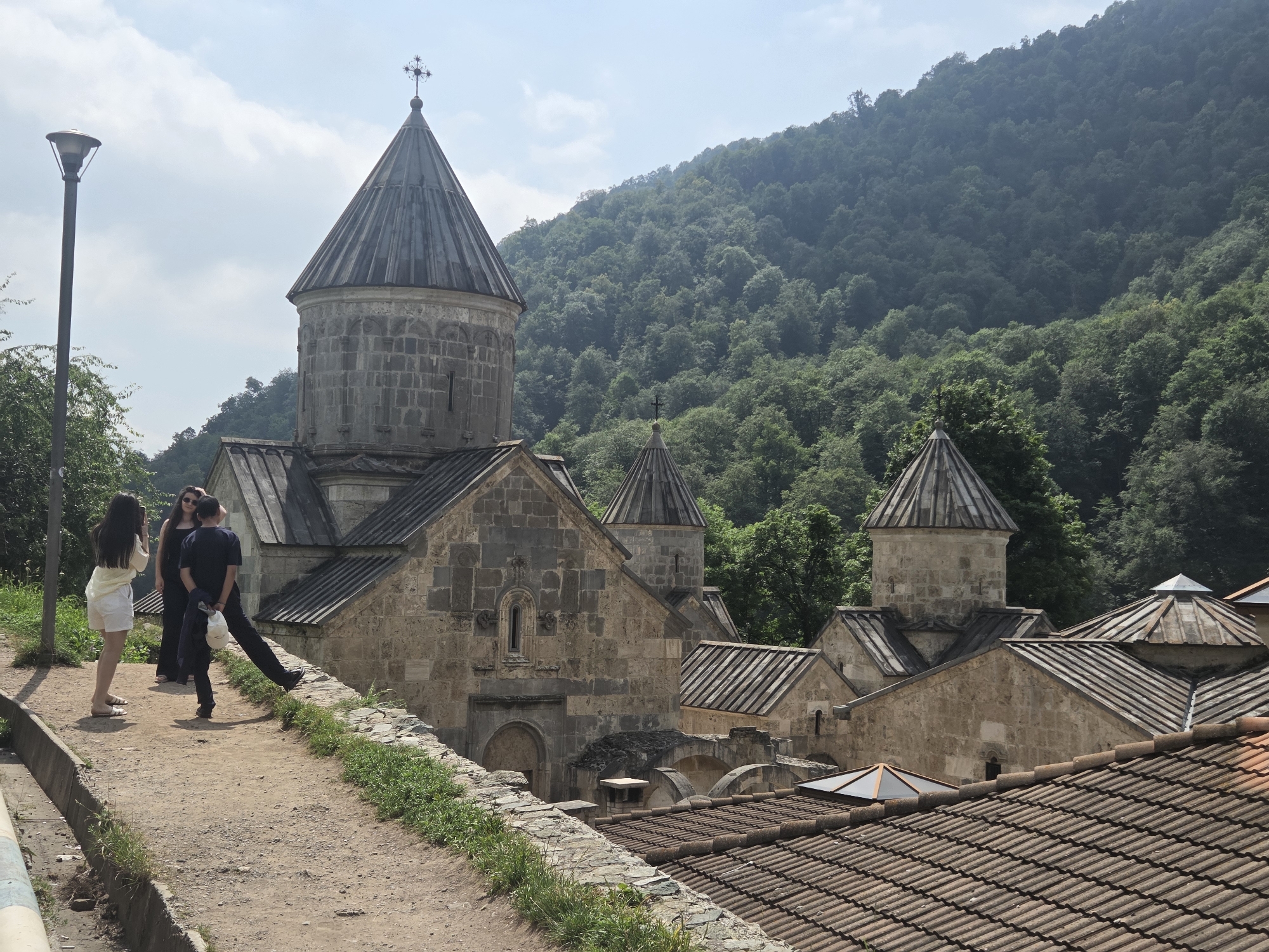 The image shows a side view of the Haghartsin Monastery complex in the Teghut region of Armenia. The monastery is constructed from stone and features multiple domed structures topped with crosses. In the foreground, a dirt path runs alongside a stone wall covered in grass. Three individuals are visible on the path: two women and one man. One woman in a white dress and shorts stands with her back to the camera, looking towards the monastery. The other woman, in a dark dress, stands next to her, also looking towards the monastery. The man, wearing a dark t-shirt and pants, stands with his back to the camera, facing away from the women and towards the monastery. He is holding a light-colored jacket or bag in his hand. A lamppost stands to the left of the path. Behind the monastery, a densely forested mountain slope rises under a clear sky with scattered clouds. The overall scene depicts a visit to a historical and religious site in a natural setting.