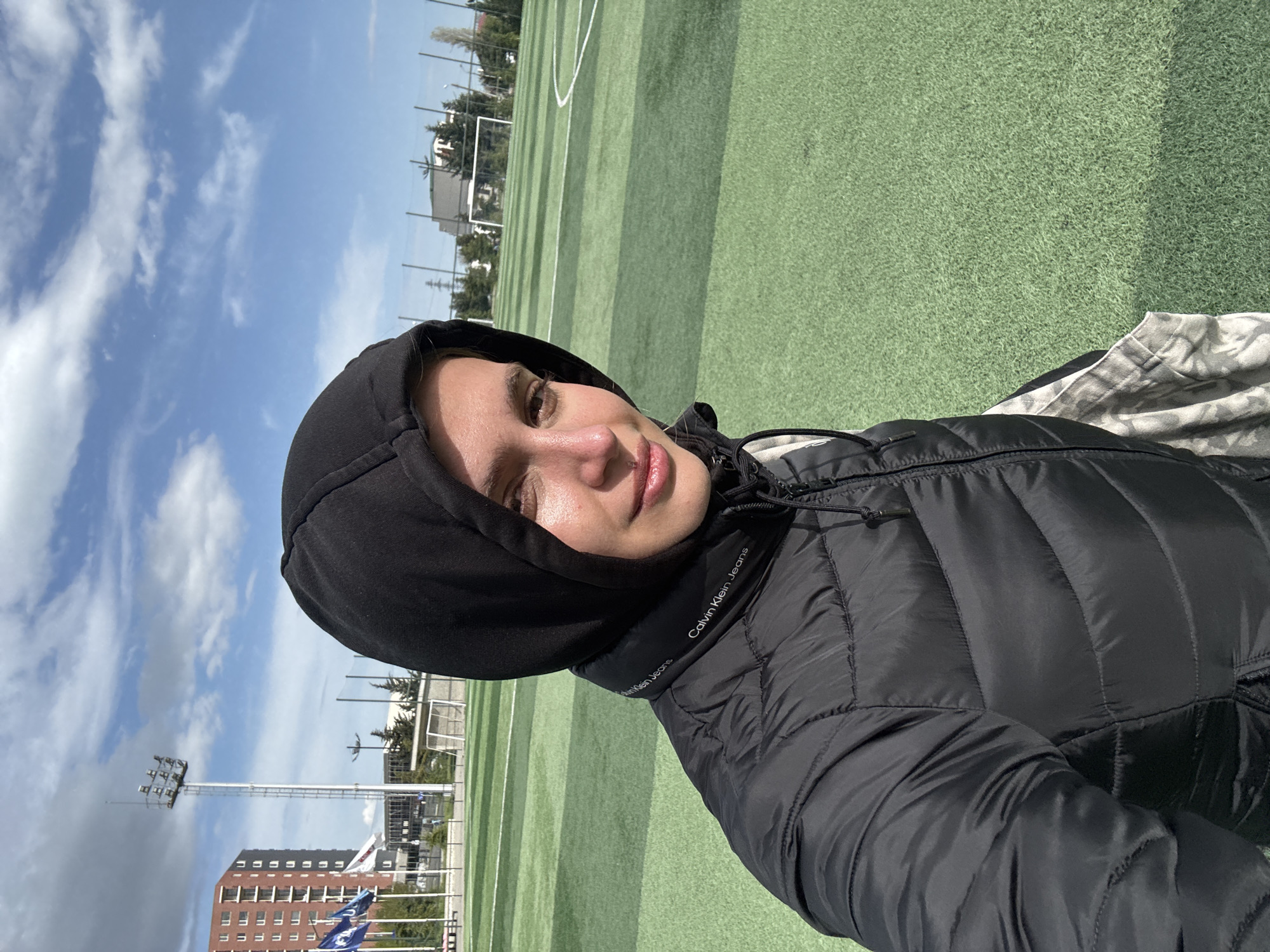 A person wearing a black Calvin Klein jacket with the hood up is taking a selfie on what appears to be a soccer field. In the background is a building, a light tower, blue and white flags, and a blue sky with clouds. The field is green. While it's impossible to determine the exact location within Tbilisi without additional information or visual cues, based on the presence of the soccer field, the type of buildings, and the light tower, it is plausible that this photo was taken at or near a sports complex or a park with sporting facilities in Tbilisi, Georgia.