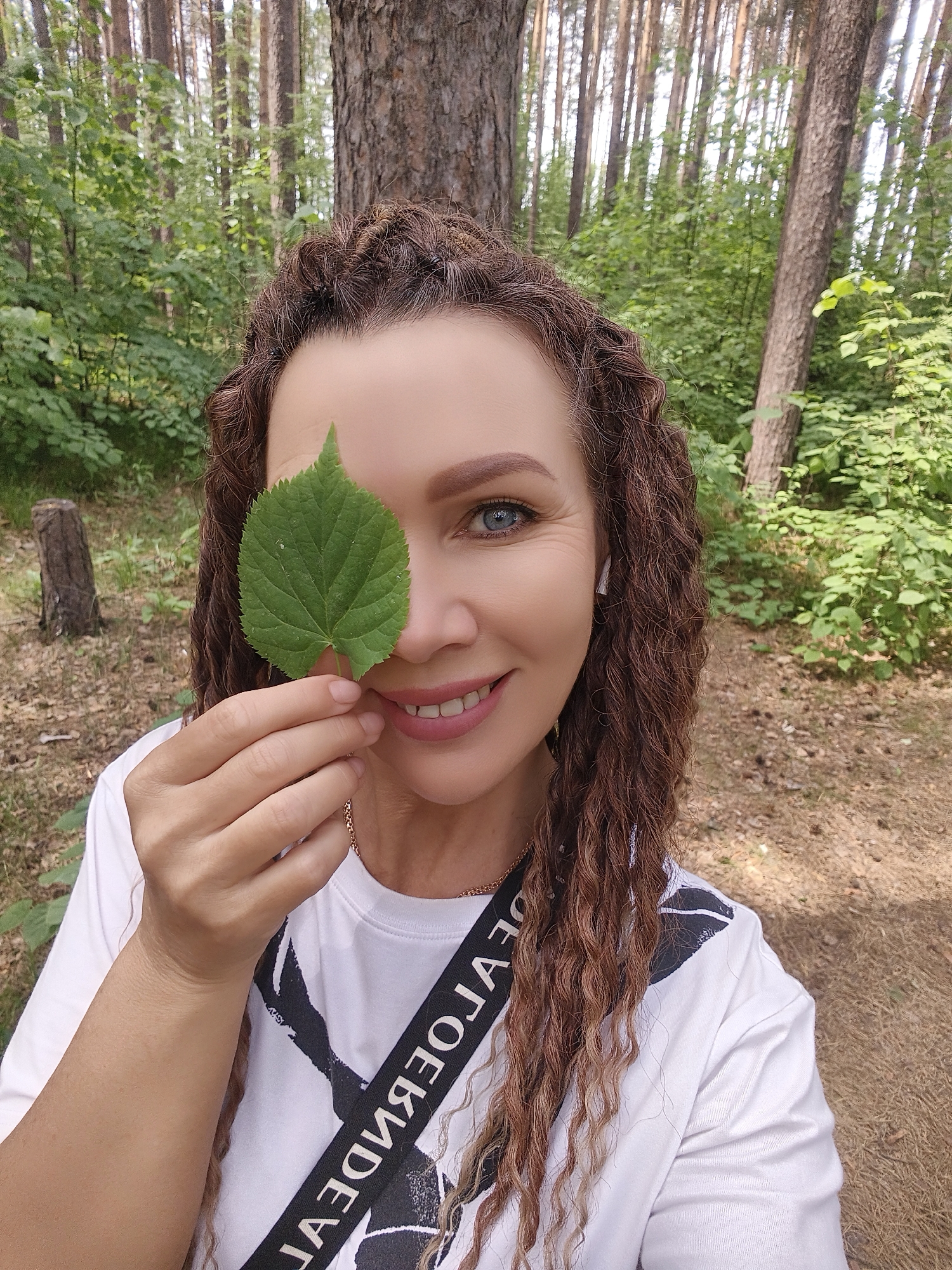A woman with curly brown hair is holding a green leaf in front of her left eye. She is smiling and has light blue eyes. She is wearing a white T-shirt with a black and white design and a black shoulder strap with white lettering. The background is a forest with tall trees and green foliage. The ground is covered in brown pine needles. The location is Unknown, Unknown.