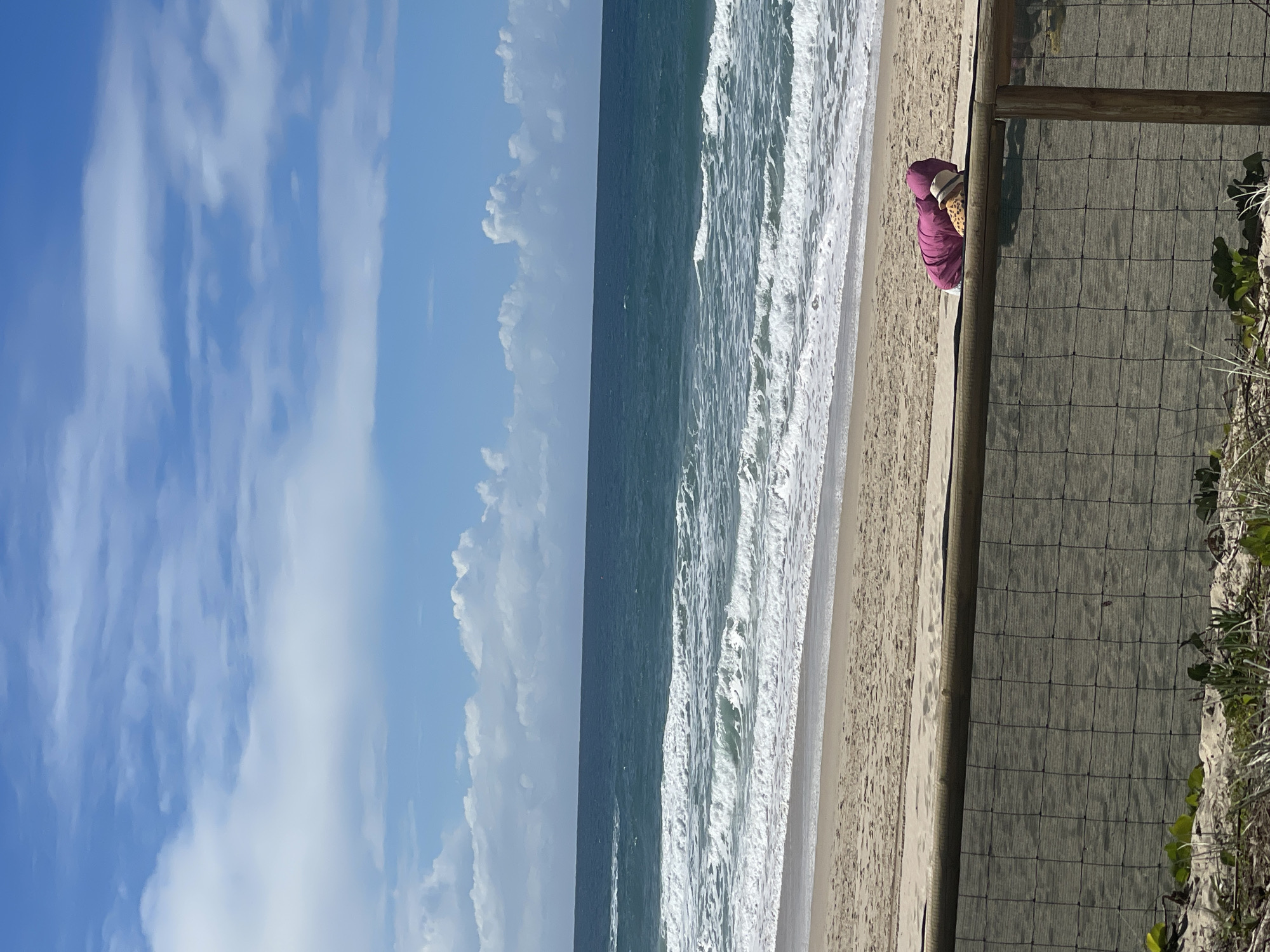 A person is sleeping on a beach with a pink blanket and a straw hat. The person is lying on their stomach facing away from the camera. The beach is sandy and there are waves crashing in the ocean. The sky is blue with white clouds. The image was taken at Surfers Paradise Beach in the Gold Coast, Australia.