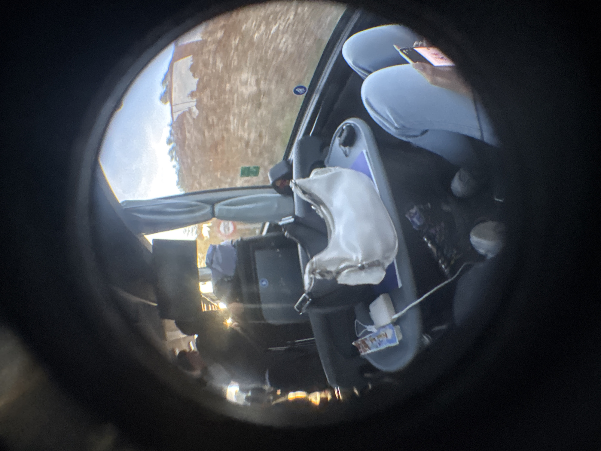 Captured through a fisheye lens, the interior of a vehicle is visible, likely a taxi, with a gear shift covered in a white protective boot. The passenger in the back is occupied with a mobile phone, wearing light blue clothing and grey shoes. The vehicle appears to be in motion, as evidenced by the blurred landscape visible through the windshield. The view shows what appears to be a street in Pozorrubielos de la Mancha, Spain.