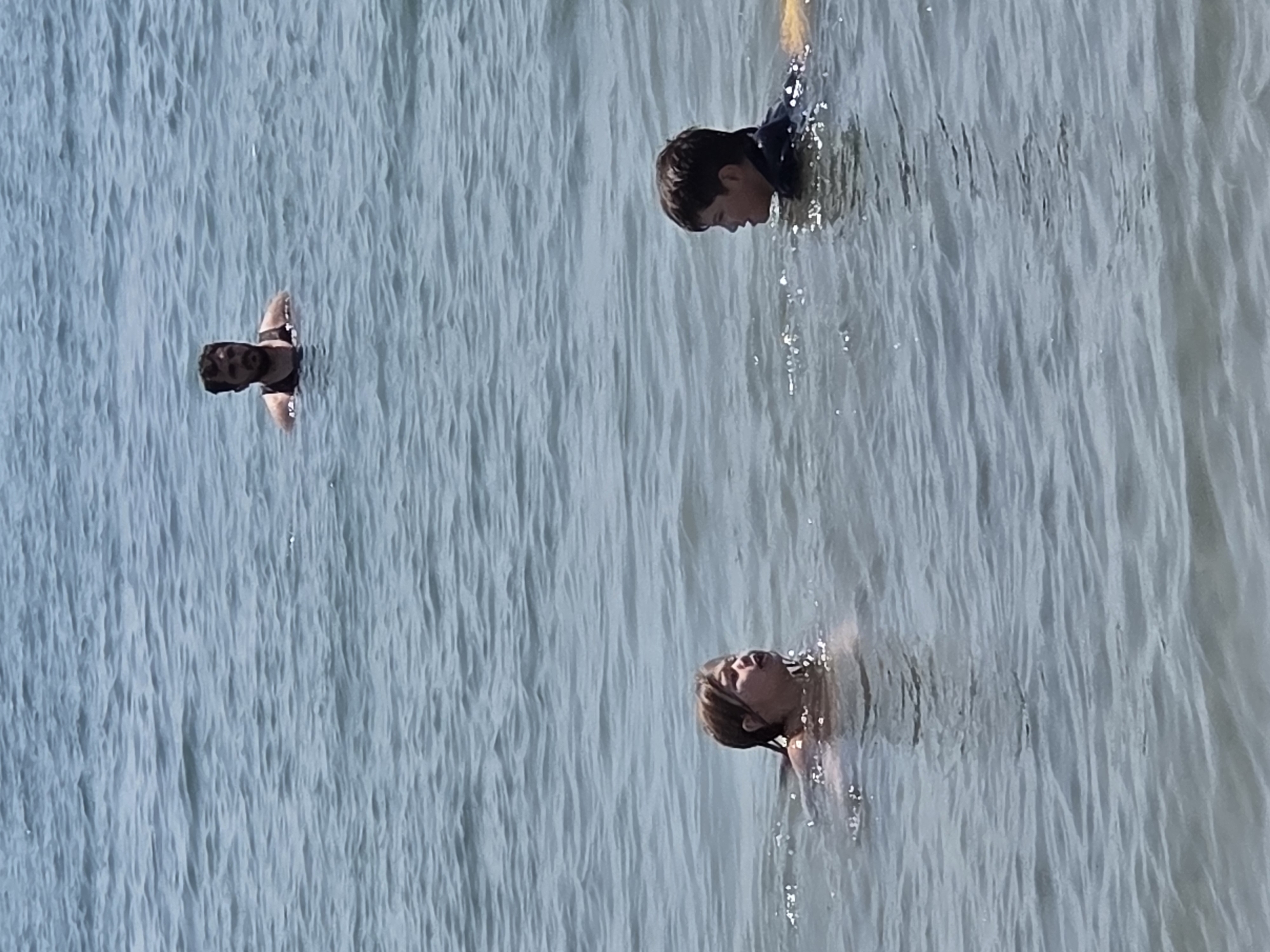 In the city of Long Beach, USA, three people are visible in the water. In the foreground, a young boy wearing a dark blue rash guard is on the right, and a young girl with blonde hair is on the left, both looking towards each other. In the background, a man with dark hair and a beard is swimming, looking forward. The water appears calm with light ripples, and the sky is bright, suggesting a sunny day.