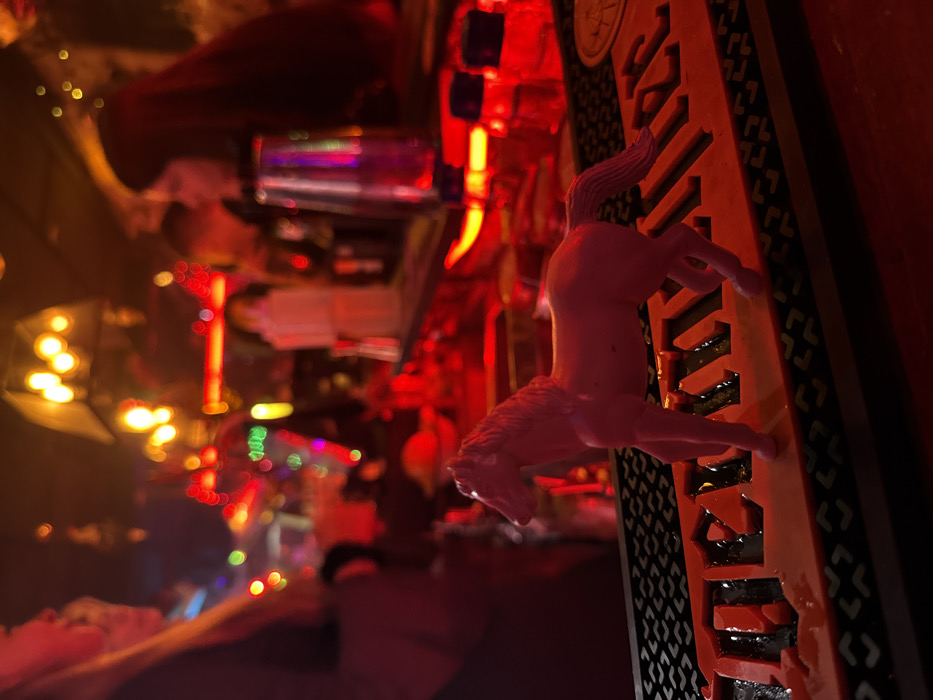 In a dimly lit, blurry setting, a plastic pink pony is perched on a decorative object. The object is tan with dark accents and a geometric pattern, possibly part of a larger structure. Red lighting dominates the scene, with other colorful lights visible in the background. The pony appears to be leaping or climbing onto the object, creating an unexpected juxtaposition in the hazy ambiance.