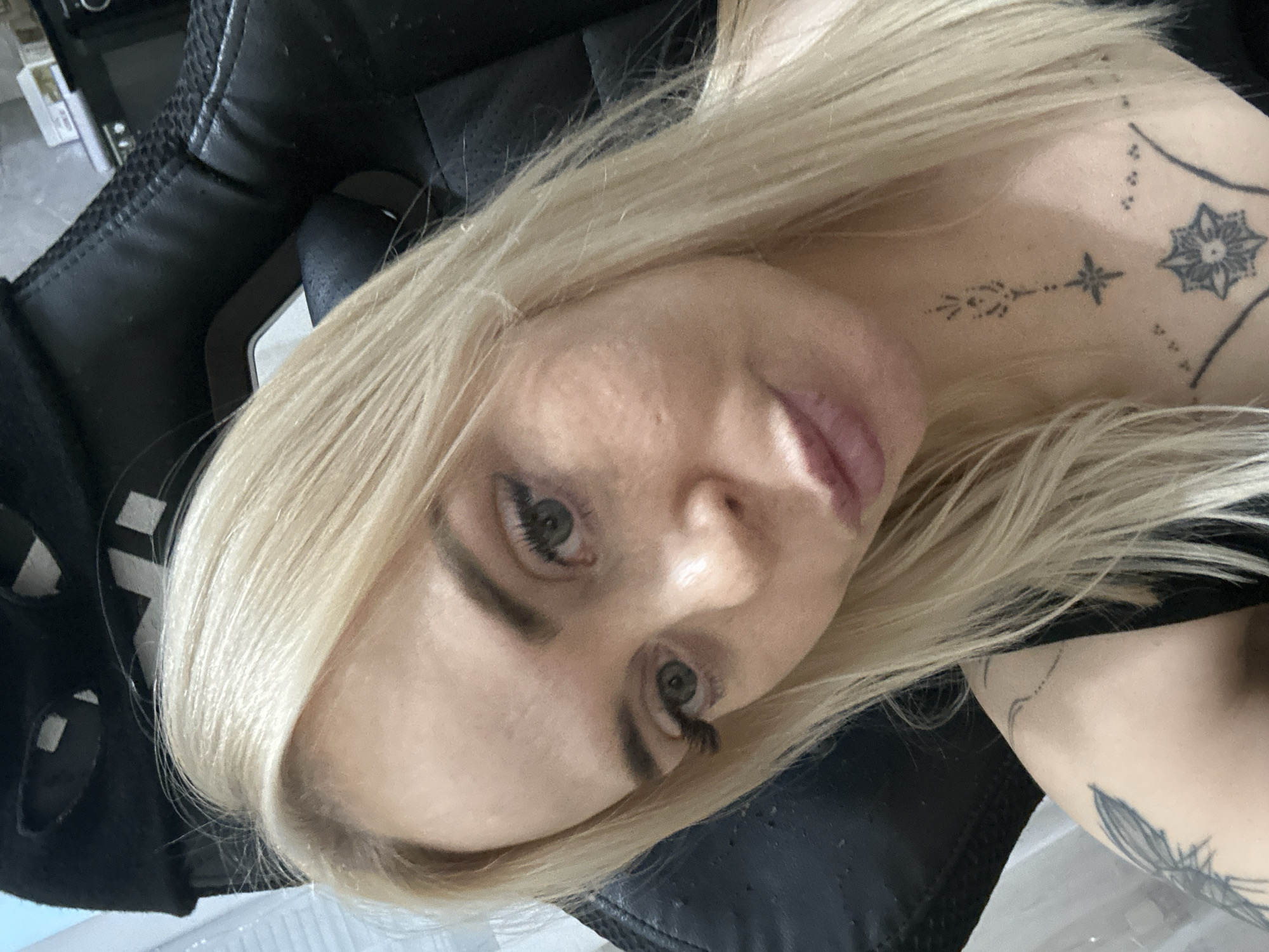 In the city of Unknown, Unknown, a fair-skinned individual with long, straight, platinum blonde hair is captured in a close-up selfie, seated indoors. The individual, identified as female, has light-colored irises, possibly greyish-blue, and looks directly at the camera with a neutral expression, lips slightly parted in a soft pout. Her eyebrows are dark and well-defined, and her eyelashes appear long, possibly enhanced. Visible facial skin texture includes minor pores and subtle blemishes.
The subject wears a black sleeveless top, with a strap visible over her left shoulder. Three distinct black ink tattoos are visible: a geometric/floral pattern on the upper chest, a smaller stylized arrow-like motif centered vertically on the throat, and a winged insect (possibly a butterfly or moth) on the upper left arm/shoulder.
She is seated in a black synthetic leather gaming or office chair, characterized by padded sections and visible stitching. The chair's headrest is partially visible behind her, showing a black strap with white square details. The background is largely out of focus, indicating a shallow depth of field. To the left, a vertical light-colored element, possibly a window blind or frame, is visible. To the right, dark, indistinct shapes suggest electronic equipment or furniture, with a hint of a silver object and a circular lens-like feature in the upper right corner.