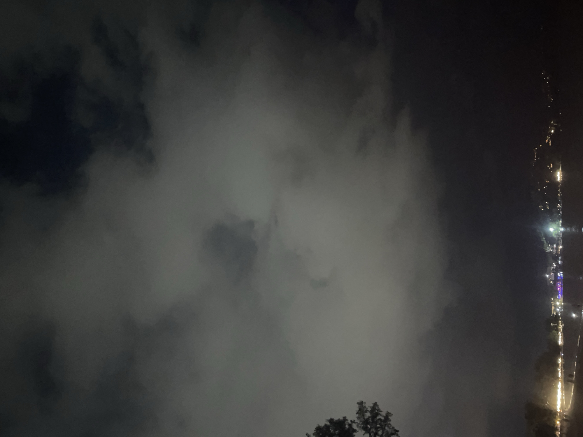 At night in Choeng Thale, Thailand, there are heavy clouds in the sky. Trees are visible at the bottom of the frame. A row of lights suggests a street or road on the right side of the image.