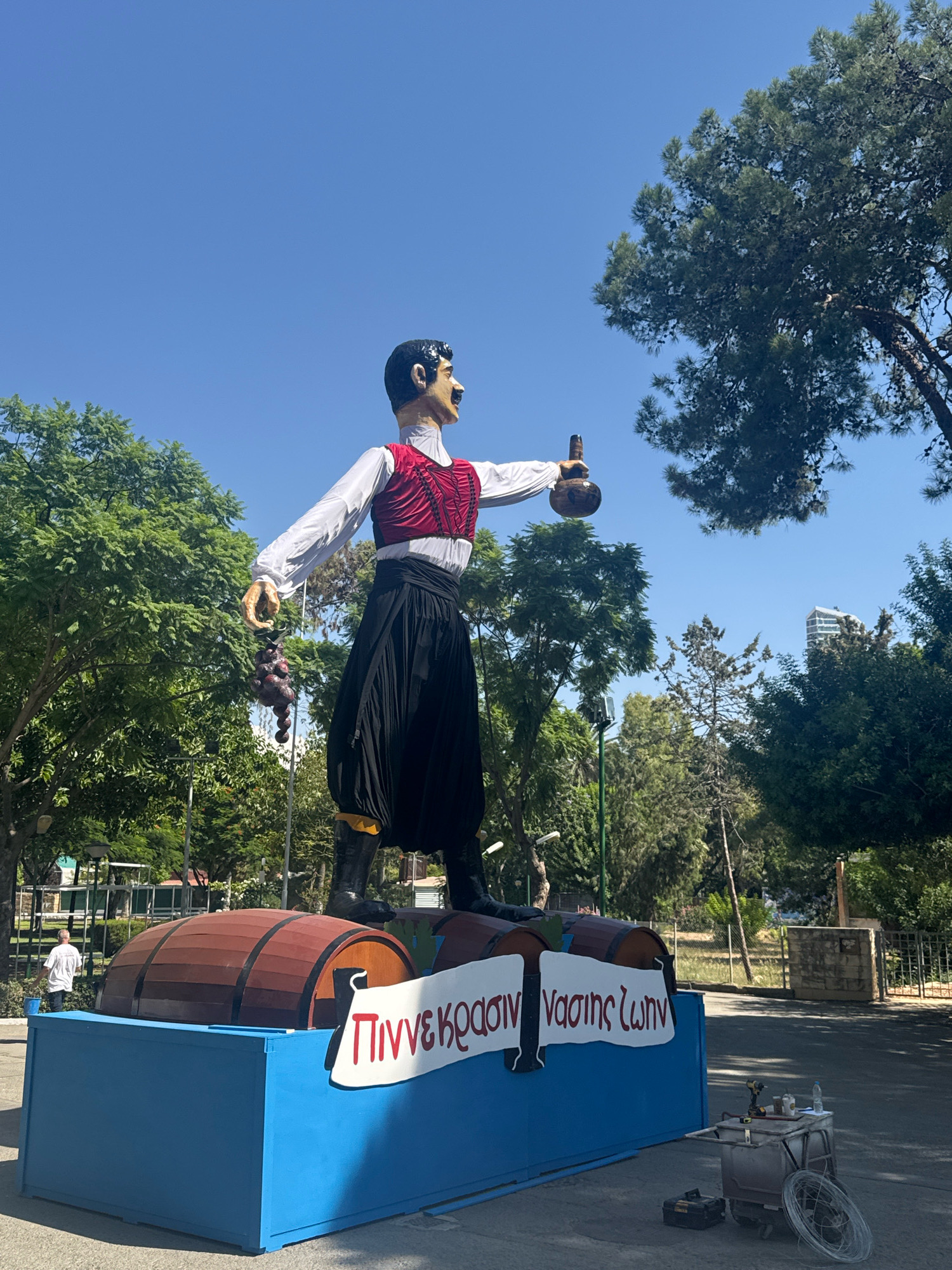 The image captures a sunny outdoor scene in Limassol, Cyprus, featuring a large, festive effigy of a man standing on a platform adorned with wooden barrels. The effigy is dressed in traditional Cypriot attire: a white long-sleeved shirt, a red vest, and a black, full skirt cinched at the waist with a sash. He holds a bunch of dark grapes in his left hand and a bottle in his right. A white banner with red lettering, appearing to be in Greek, is affixed to the front of the blue platform, reading "ΠΙΝΝΕΚΡΑΣΙΝ ΝΑΣΗΣ ΖΩΝΗ" which translates to "The Spirit of the Winemaker."
Behind the effigy, lush green trees form a vibrant backdrop under a clear blue sky. In the distance, a modern building is visible amongst the foliage, suggesting an urban environment. To the right of the platform, a cart with tools, including a drill and wire, is present, indicating ongoing or recent setup for an event. In the background, a person in a white shirt is seen near a fence, with a blue bucket in hand. The overall atmosphere suggests a cultural celebration or festival, likely related to wine production, given the effigy's attire, the grapes, the barrels, and the banner.