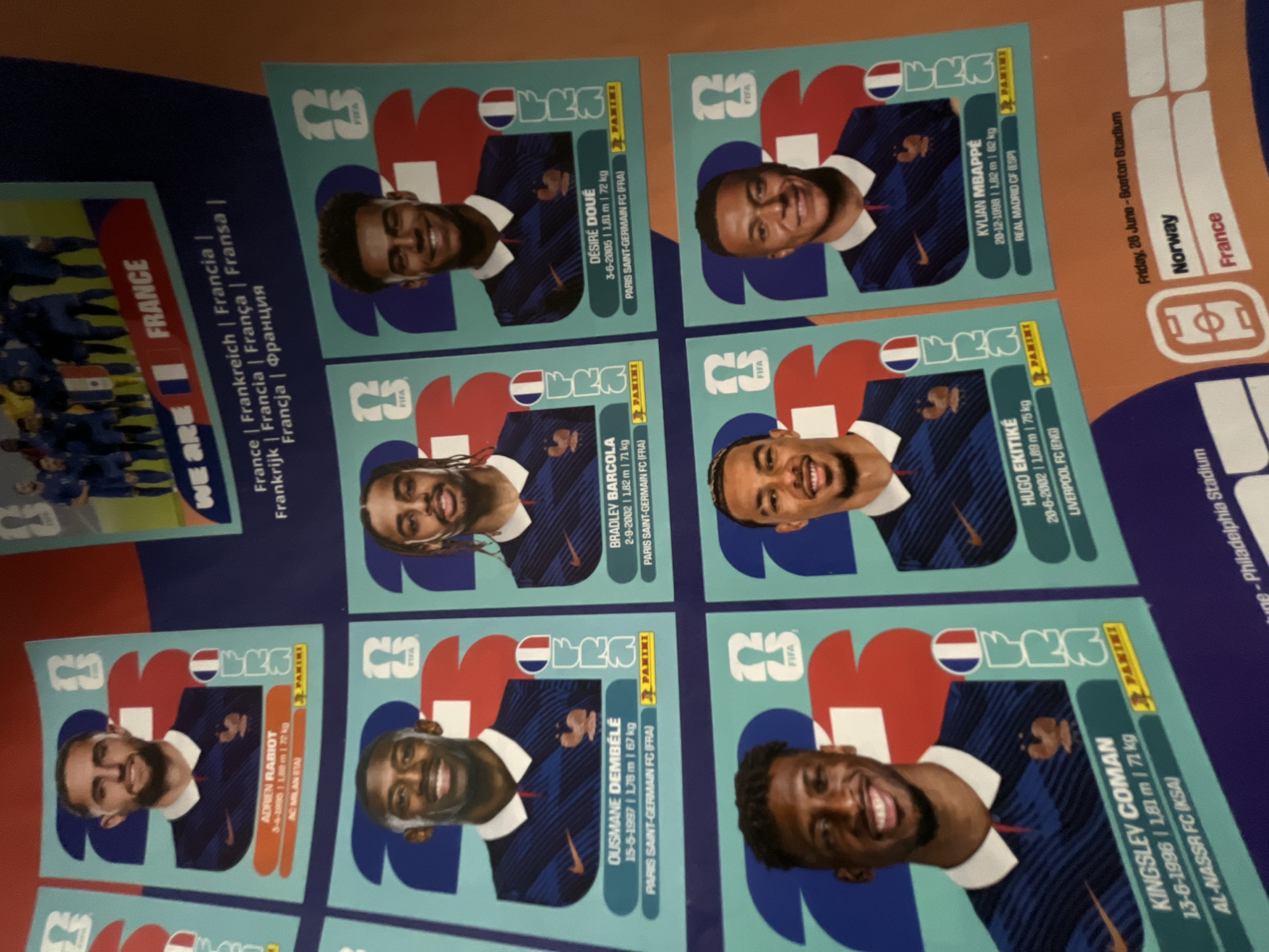 The image shows a collection of France national football team Panini stickers. Six player stickers are visible: Adrien Rabiot, Ousmane Dembélé, Kingsley Coman, Bradley Barcola, Désiré Doué, Kylian Mbappé, and Hugo Ekitiké. In the bottom right corner, there is a section of a schedule indicating a match between Norway and France on Friday, June 26th at Boston Stadium. Another visible stadium name is "Philadelphia Stadium". The image composition appears to be a somewhat cluttered arrangement of items on a surface.