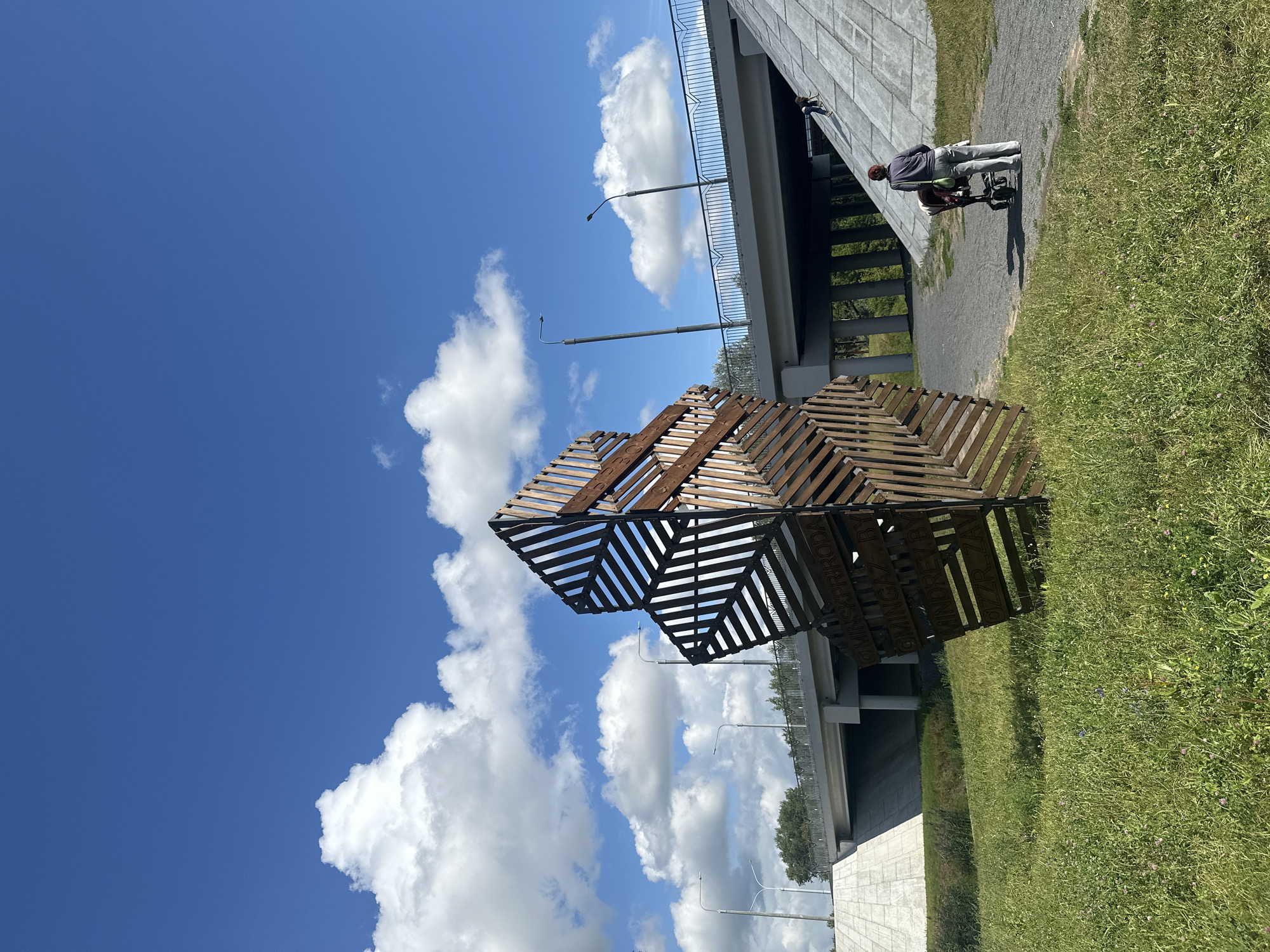 The image depicts an outdoor scene in Minsk, Belarus, under a bright blue sky with scattered white clouds. In the foreground, a large, abstract wooden sculpture made of interconnected geometric shapes stands on a grassy area. The sculpture has text inscribed on it, partially visible. To the right of the sculpture, a paved path runs alongside a grassy slope. A person is pushing a stroller on the path, facing away from the camera. Further in the background, a modern concrete bridge spans over a road, with streetlights visible on the bridge. Another person is seen climbing the concrete slope to the right of the bridge. The environment appears to be a park or recreational area near a transportation infrastructure.