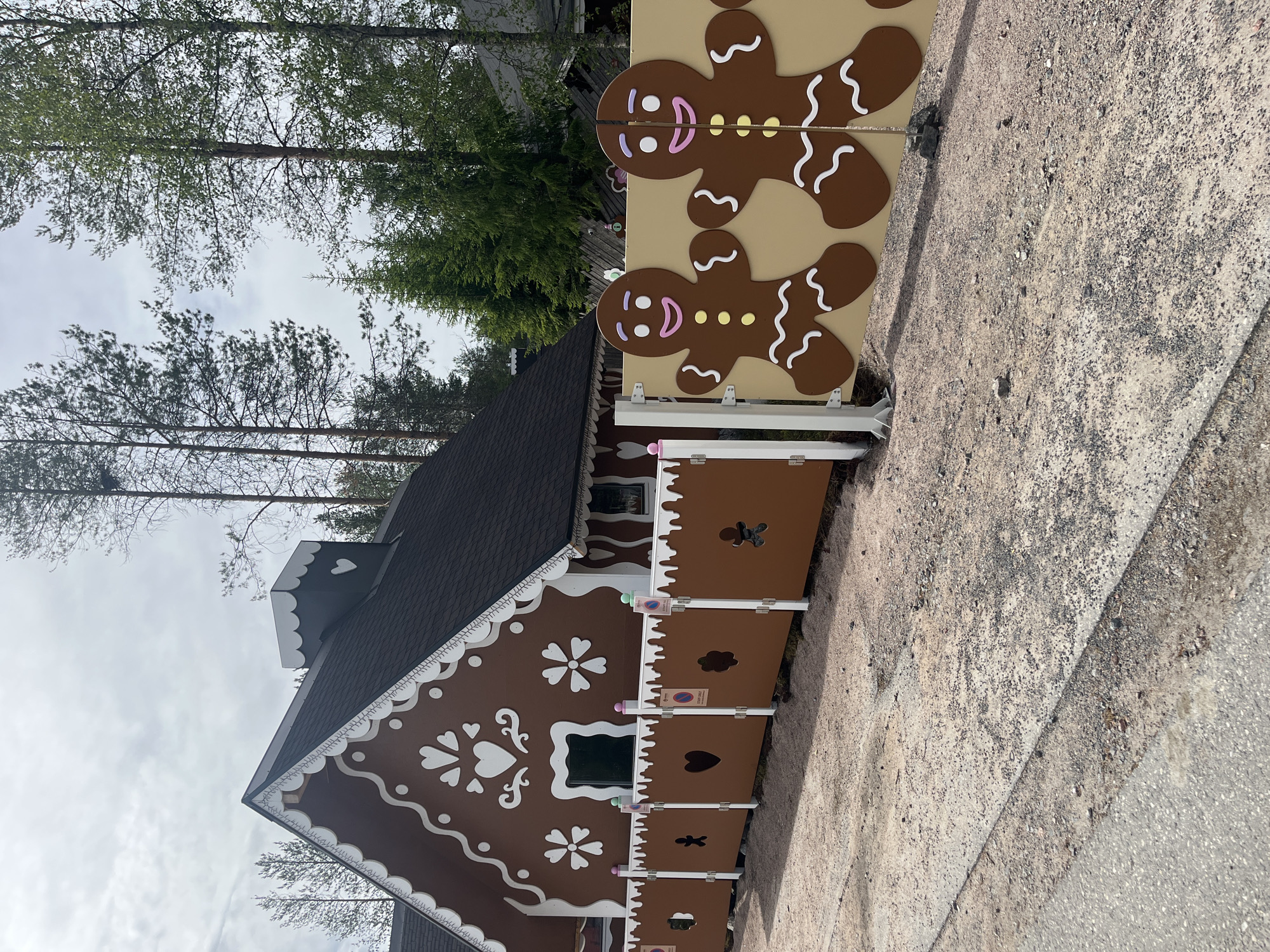 A gingerbread-themed house with brown siding and white trim, featuring heart and flower decorations, is visible in Rovaniemi, Finland. A decorative fence in front of the house is adorned with cut-out shapes like hearts, gingerbread men, and flowers. Two large, cartoonish gingerbread men figures stand on a beige panel to the right of the fence. The scene is set against a backdrop of tall evergreen and birch trees under a cloudy sky. The ground in front of the fence is a mixture of gravel and pavement.