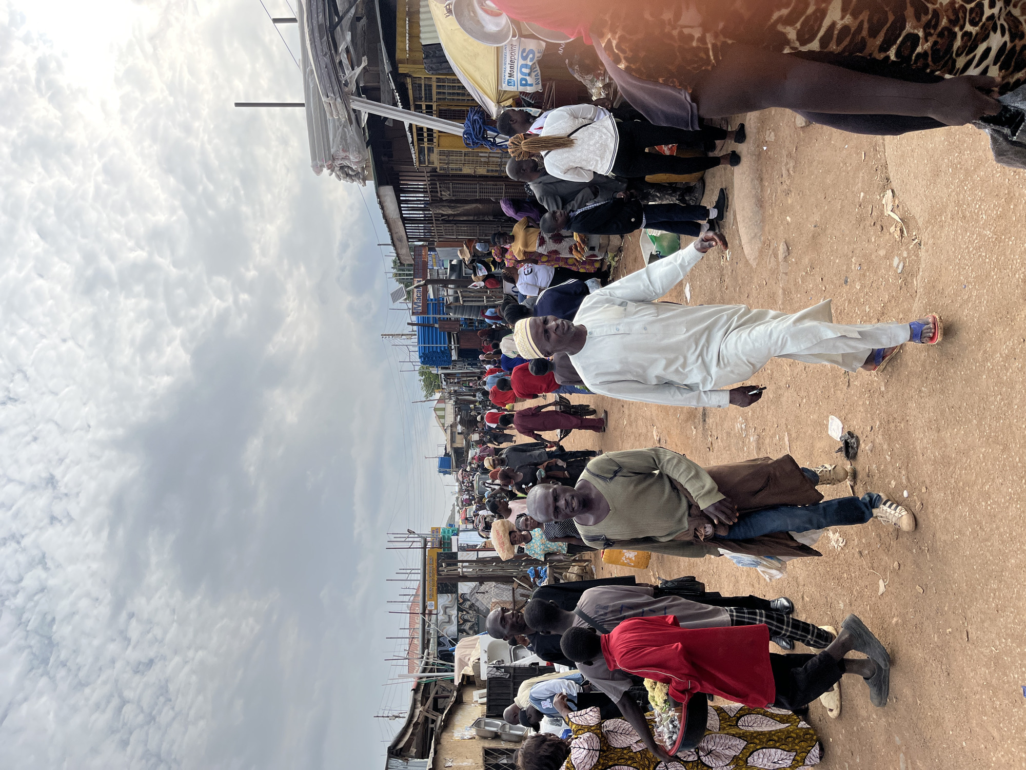 The image depicts a bustling outdoor market scene in Gura Top, Nigeria. A diverse crowd of people are gathered, many of whom are walking, standing, or looking at their surroundings. The setting appears to be a street lined with temporary structures and stalls, suggesting a commercial area. The ground is a mix of dirt and paved surfaces.
The time of day seems to be daytime, given the illumination, and the weather appears to be overcast with a cloudy sky. The people are dressed in various casual and traditional clothing. Some individuals are carrying items, and there's a sense of everyday activity, with a focus on commerce and social interaction.
Notable details include the presence of several individuals in traditional Nigerian attire, as well as more modern clothing. There is a concentration of people in the mid-ground, some of whom appear to be engaged in conversation or browsing. The overall atmosphere suggests a lively and active community marketplace.
Visible text in the image includes signage on stalls and some labels on products, though much of it is difficult to decipher due to the image quality and perspective. One sign prominently features "Montepoint" and "POS AIRTIME".