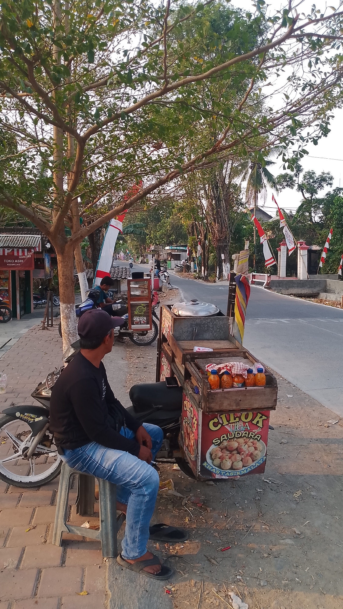 In Karanglangu, Indonesia, a man wearing a black long-sleeved shirt, blue jeans, and a dark baseball cap sits on a gray plastic stool facing away from the camera. He is seated next to a motorcycle and a wooden food cart. The food cart has a sign that reads "CILOK TIGA SAUDARA" and displays images of food items. Several bottles of sauce are visible on top of the cart. The motorcycle is parked on a paved area next to a dirt path. In the background, a paved road stretches into the distance, lined with trees and buildings. Red and white flags are visible along the road. The scene appears to be outdoors during daylight hours, with warm sunlight casting shadows.