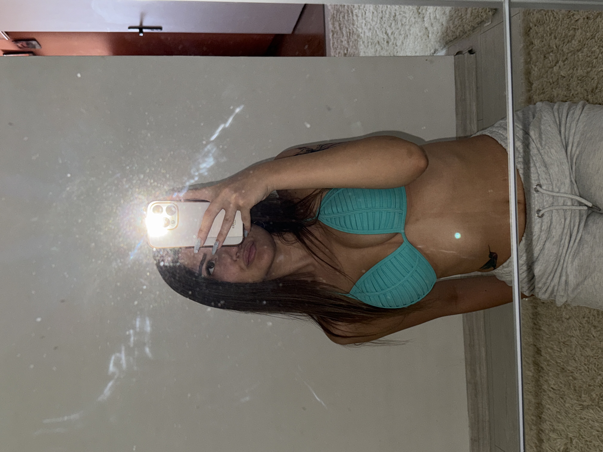 In an indoor setting, a young woman with long, dark hair is captured in a mirror selfie. She is wearing a turquoise halter-neck bikini top and light grey sweatpants. Her right hand holds a smartphone, positioned to take the picture. The background consists of a light-colored wall, a mirror, and a beige carpeted floor. A doorway with a dark-colored frame is visible to the right, along with a partially visible white door. The image appears to be taken in a residential environment. There are no discernable landmarks or elements within the image that would indicate a specific location in Sofia, Bulgaria.