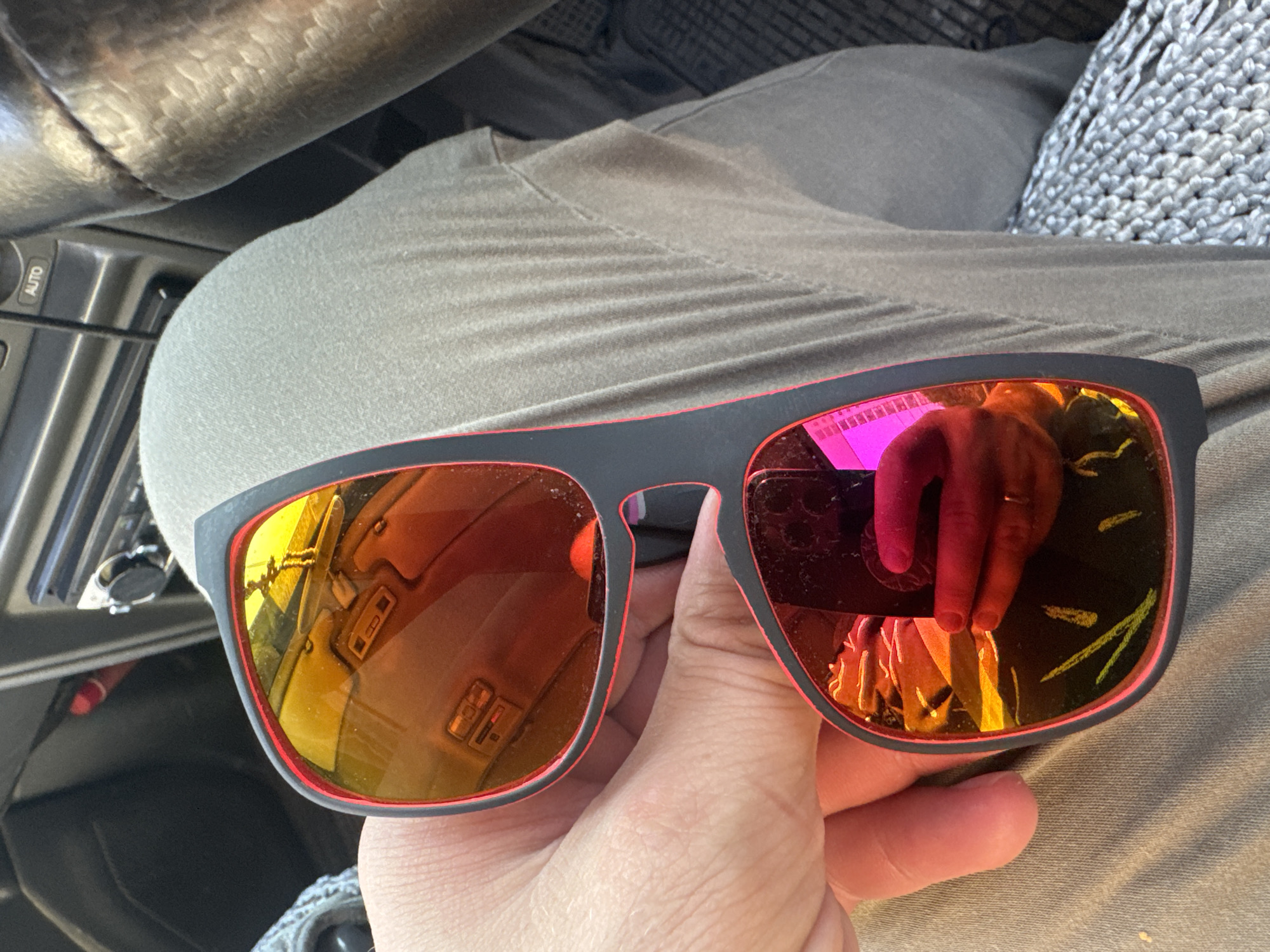 A person is holding a pair of black sunglasses with red trim and reflective orange lenses. The lenses reflect the interior of a vehicle, including a dashboard with buttons and a steering wheel. The person's hand and fingers are visible, and they appear to be wearing rings. The background shows a gray textile fabric, likely clothing. The image does not provide any specific visual clues to determine the location within Almaty, Kazakhstan.
