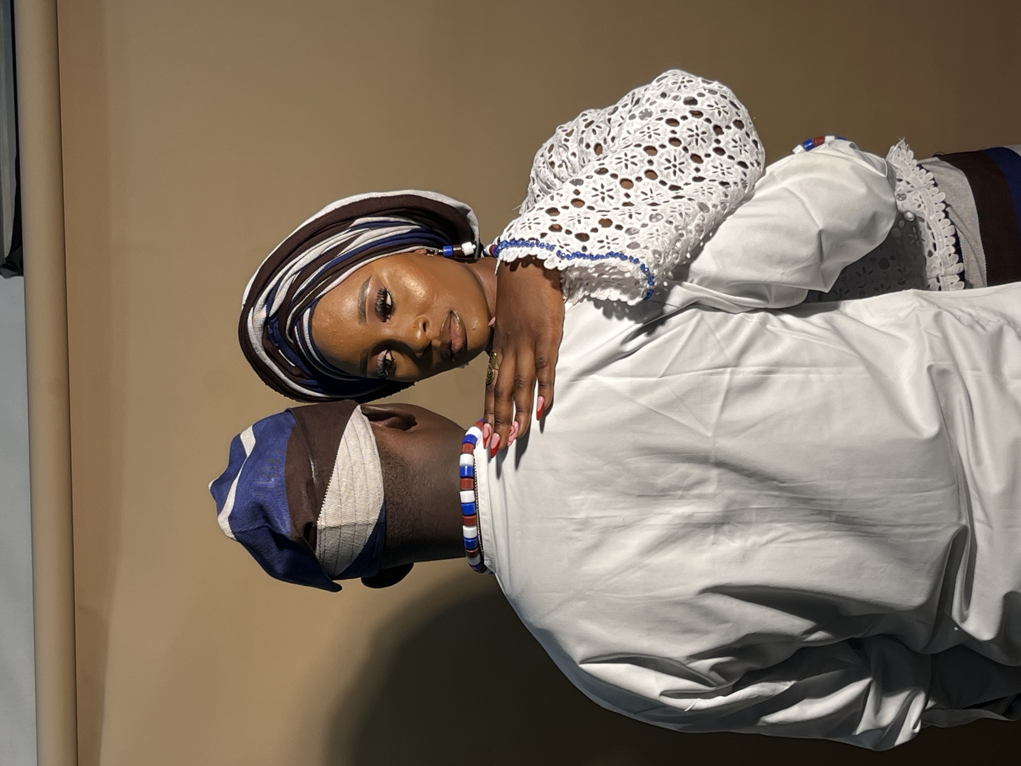 The image depicts a man and a woman from behind, embracing. The man wears a white long-sleeved shirt and a patterned cap. The woman is adorned in a white lacy top with blue bead accents and a matching headwrap. Her hand is placed on the man's shoulder. The background is a plain, light brown wall. The scene is lit with warm, directional light casting shadows. There is no indication of the specific location within Gura Top, Nigeria.