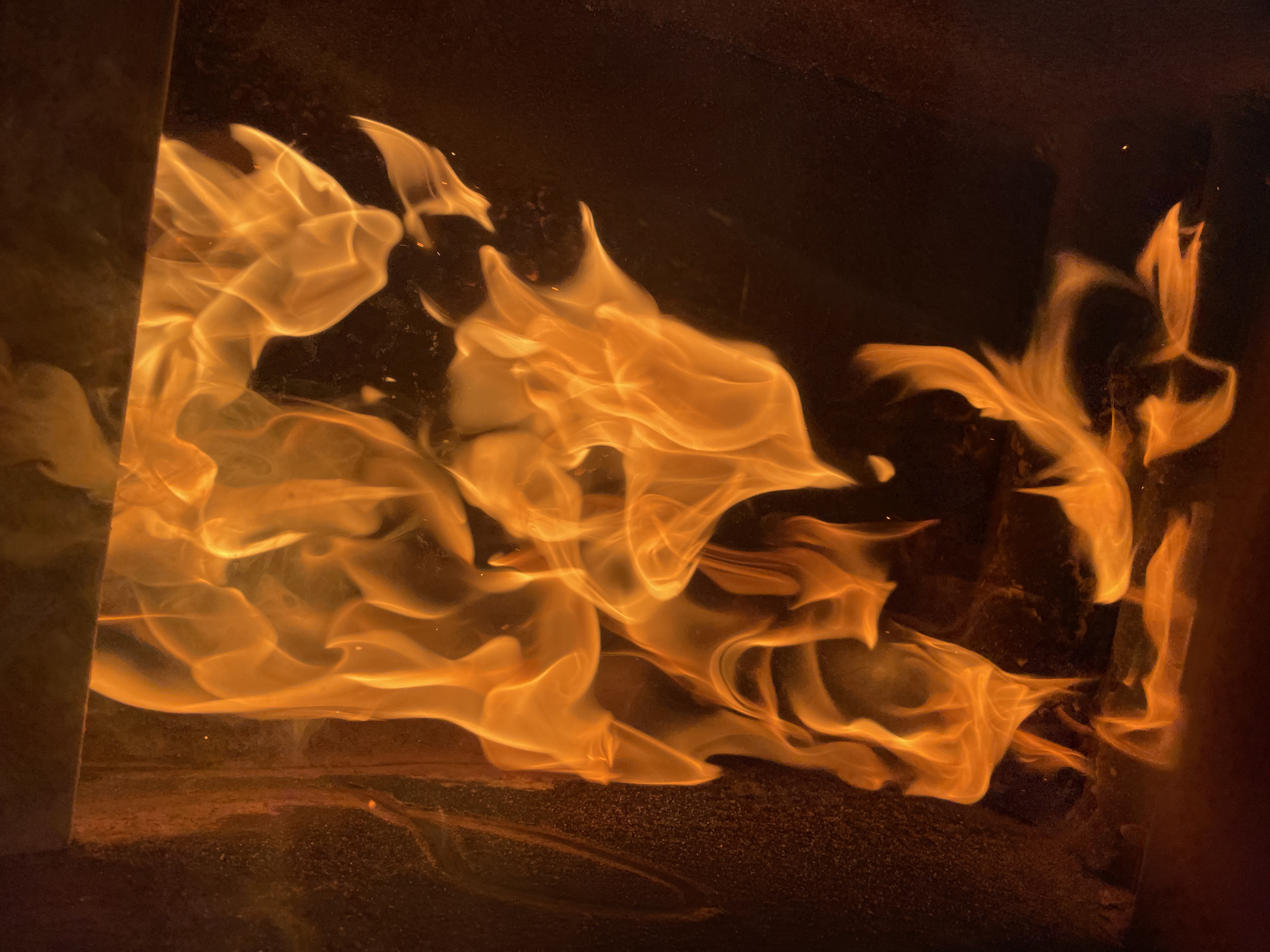 The image shows flames inside a fireplace or wood-burning stove. The fire is burning with an orange and yellow hue. The flames are contained within a dark enclosure. The bottom of the enclosure is covered in dark ashes. No people are visible. There is no information suggesting it is located in the city of Zlatograd, Bulgaria.