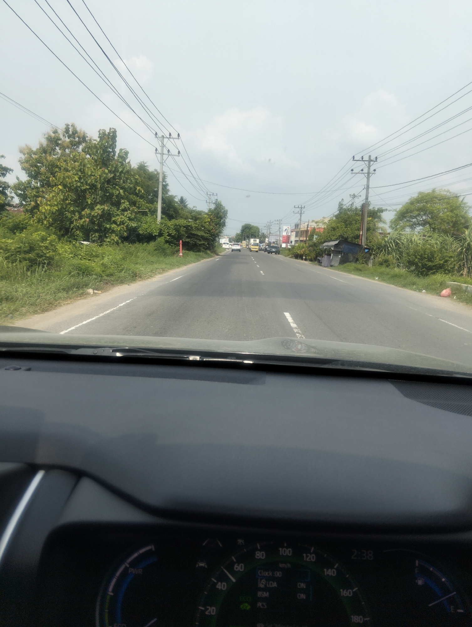 The image depicts a forward-facing view from the interior of a vehicle driving on a two-lane asphalt road. The road is flanked by lush green vegetation and trees. Utility poles with multiple wires are visible along the side of the road. In the distance, there are buildings and other vehicles, including a truck and a few cars. The sky is overcast with a pale, hazy appearance. The dashboard of the vehicle is partially visible in the foreground, displaying a speedometer and other indicators. The dashboard shows the time as 2:38 and various labels such as "PWR", "ECO", "LDA", "BSM", and "PCS". The setting appears to be a semi-rural or suburban area on a cloudy day.